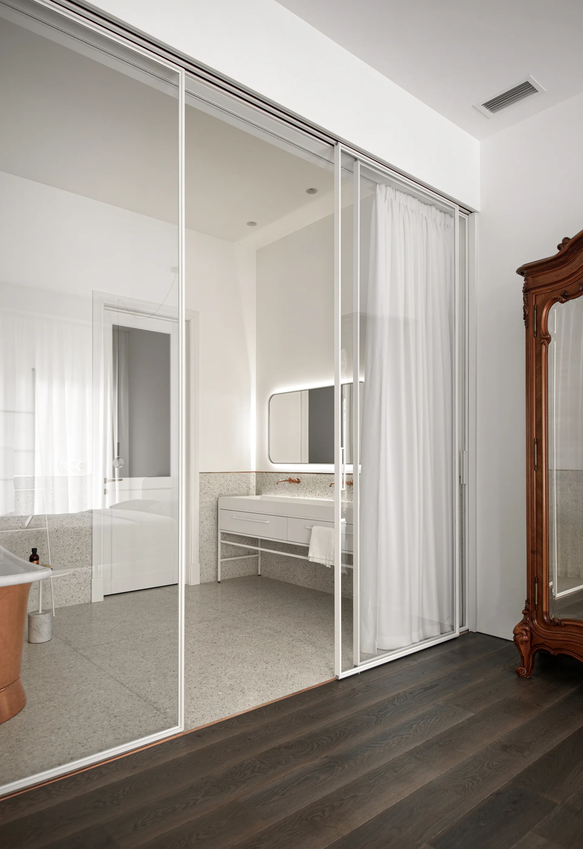 Large glass sliding doors separate a bedroom with dark wood floors from a minimalist bathroom with gray terrazzo tiles, a white vanity, and a backlit mirror.