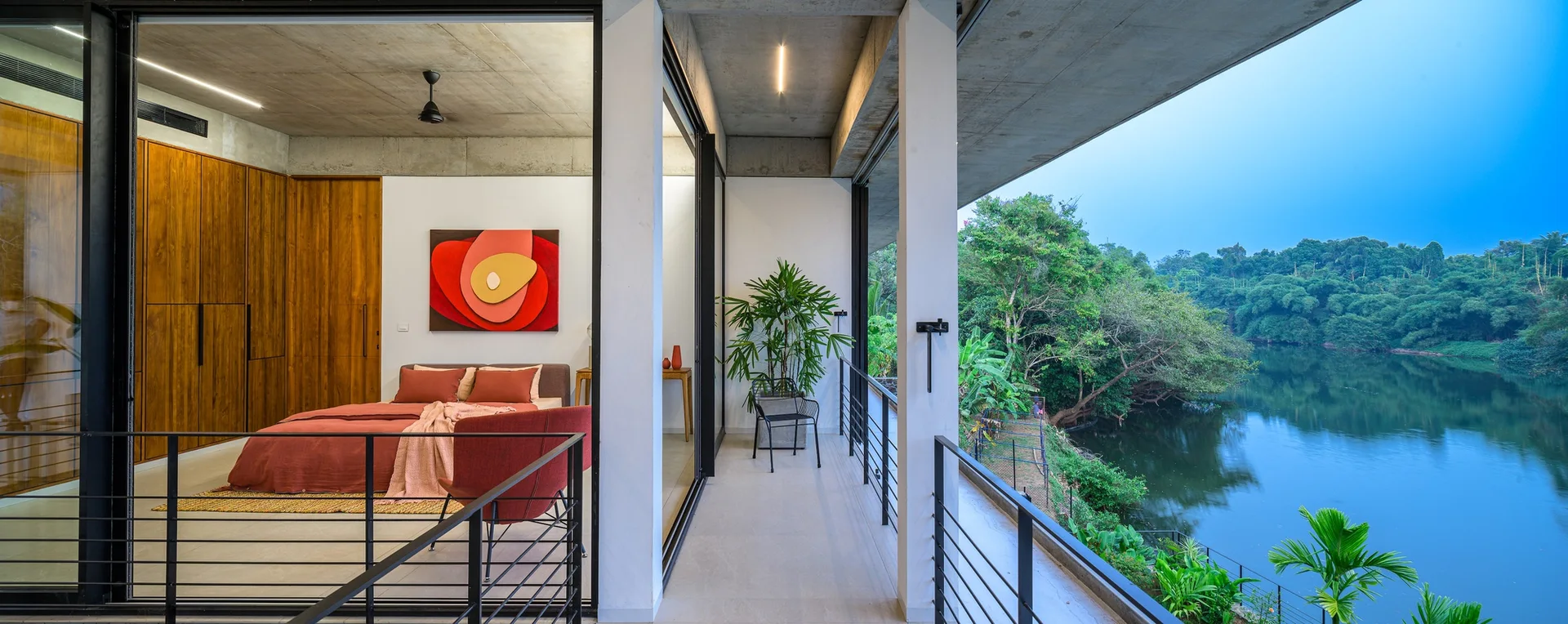 A modern bedroom with a concrete ceiling, wood wardrobes, and terracotta bedding opens onto a balcony overlooking a river.