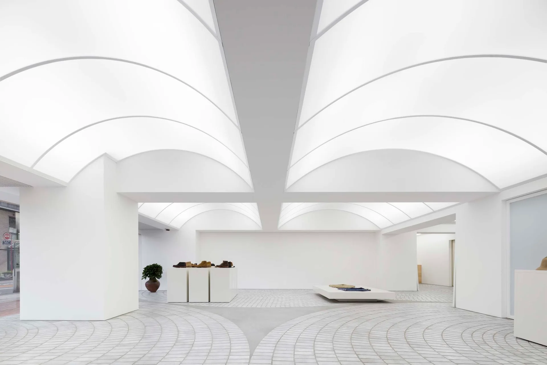 A minimalist showroom features a glowing vaulted ceiling, white display plinths, and light grey floor tiles arranged in a curved pattern.