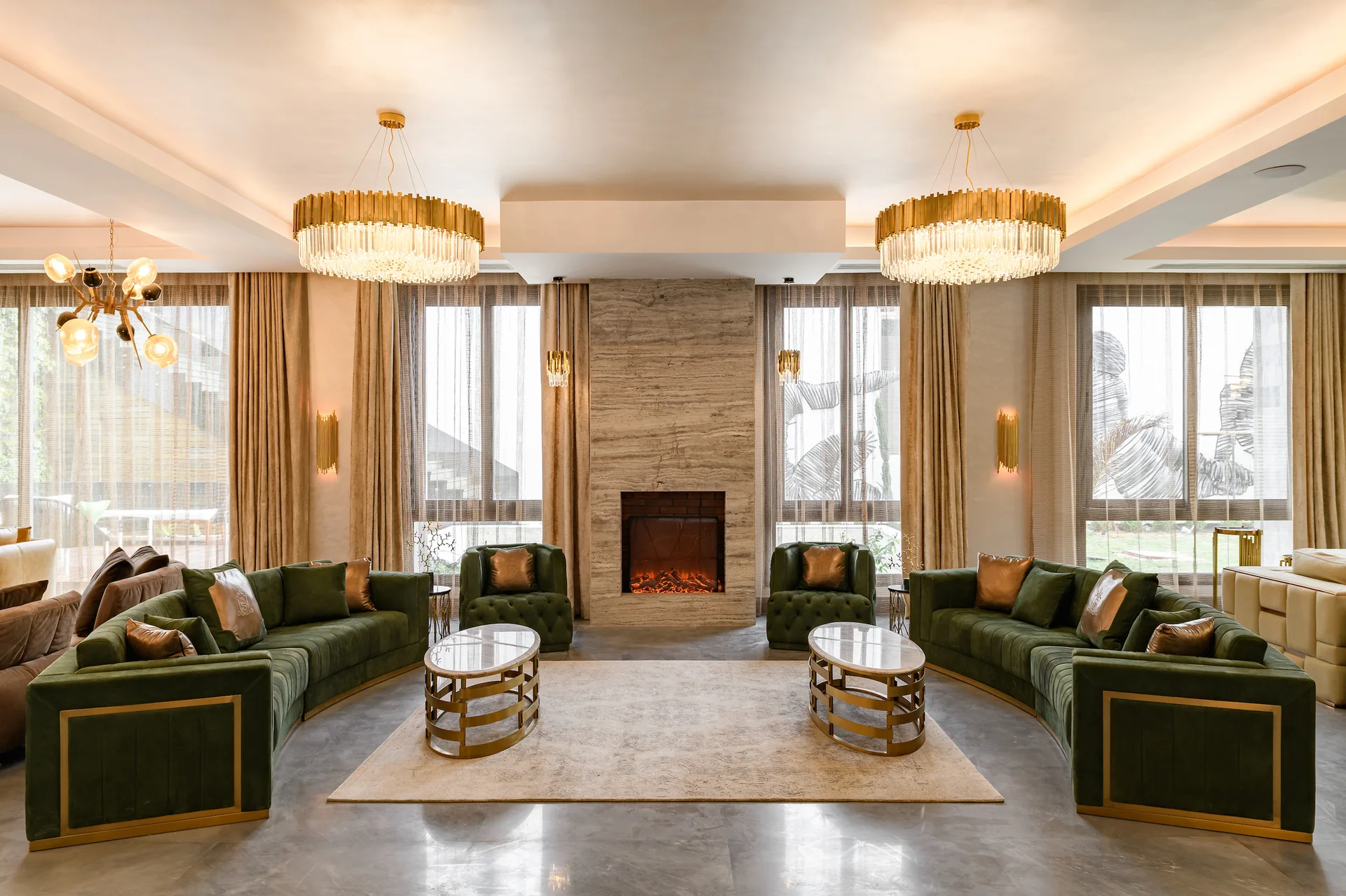 A symmetrical living room features two green velvet sofas, matching armchairs, a central stone fireplace, and gold circular chandeliers.