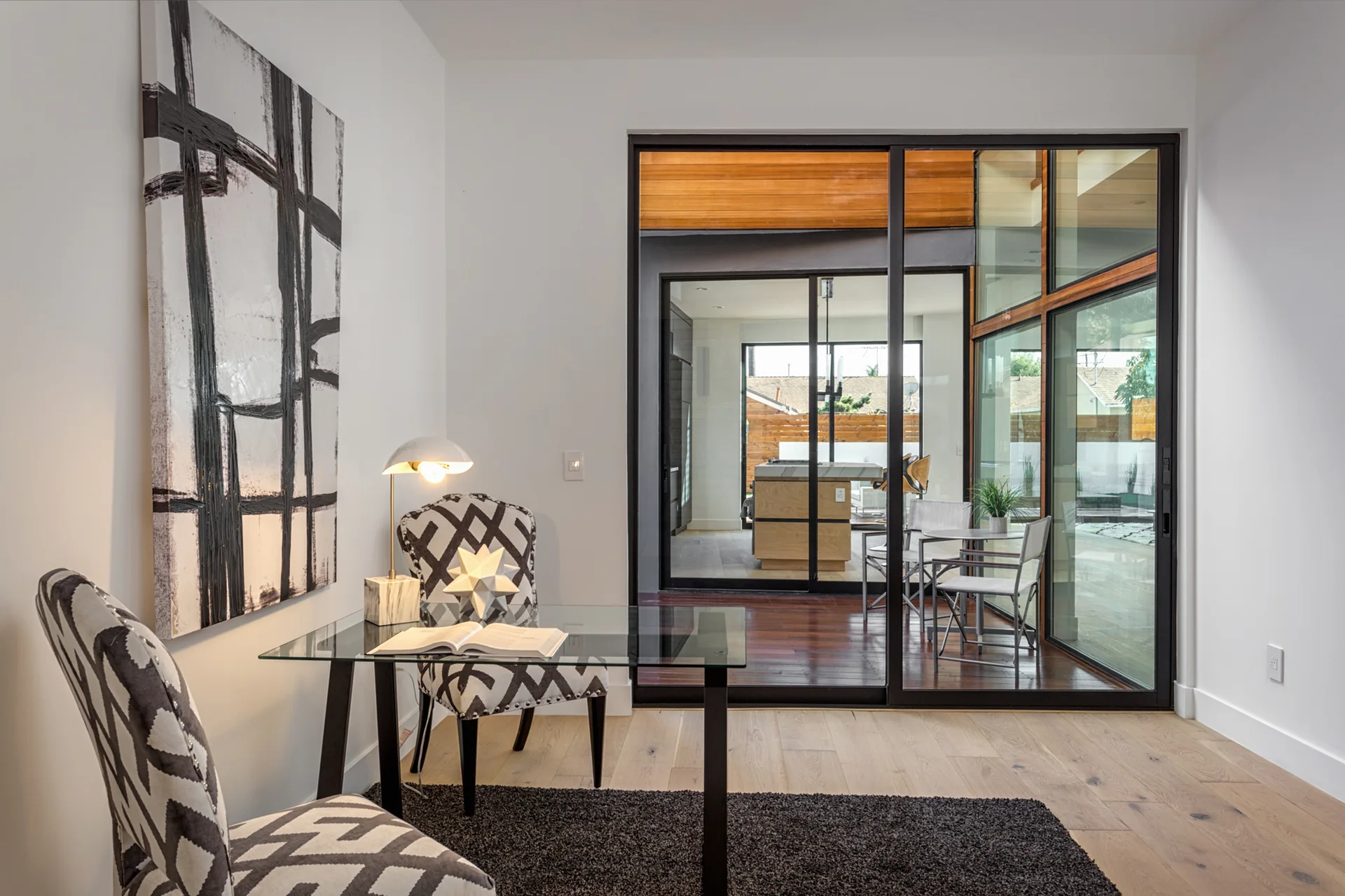 A modern home office features a glass-top desk, geometric upholstered chairs, and a large black-framed sliding glass door.
