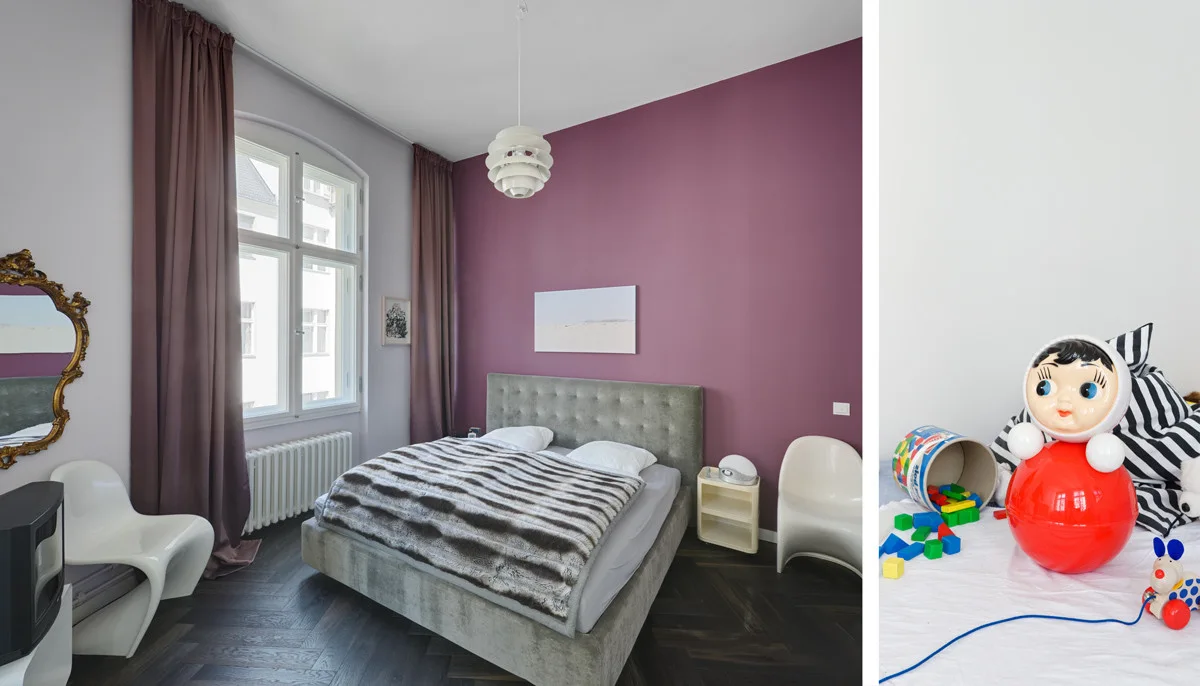 A bedroom features a plum-colored accent wall behind a grey upholstered bed, dark wood herringbone flooring, and mauve drapes beside a tall arched window.