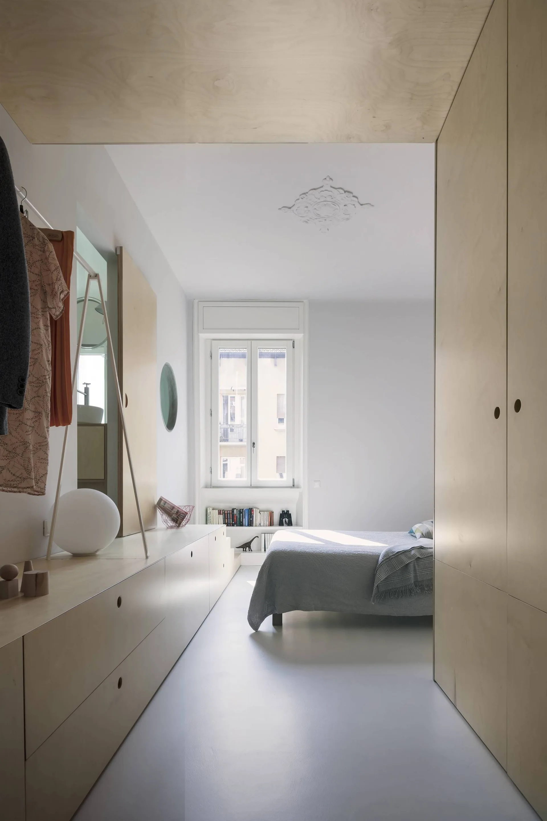 A minimalist bedroom features light plywood cabinets, a grey bed, and a white ceiling with an ornate plaster medallion.