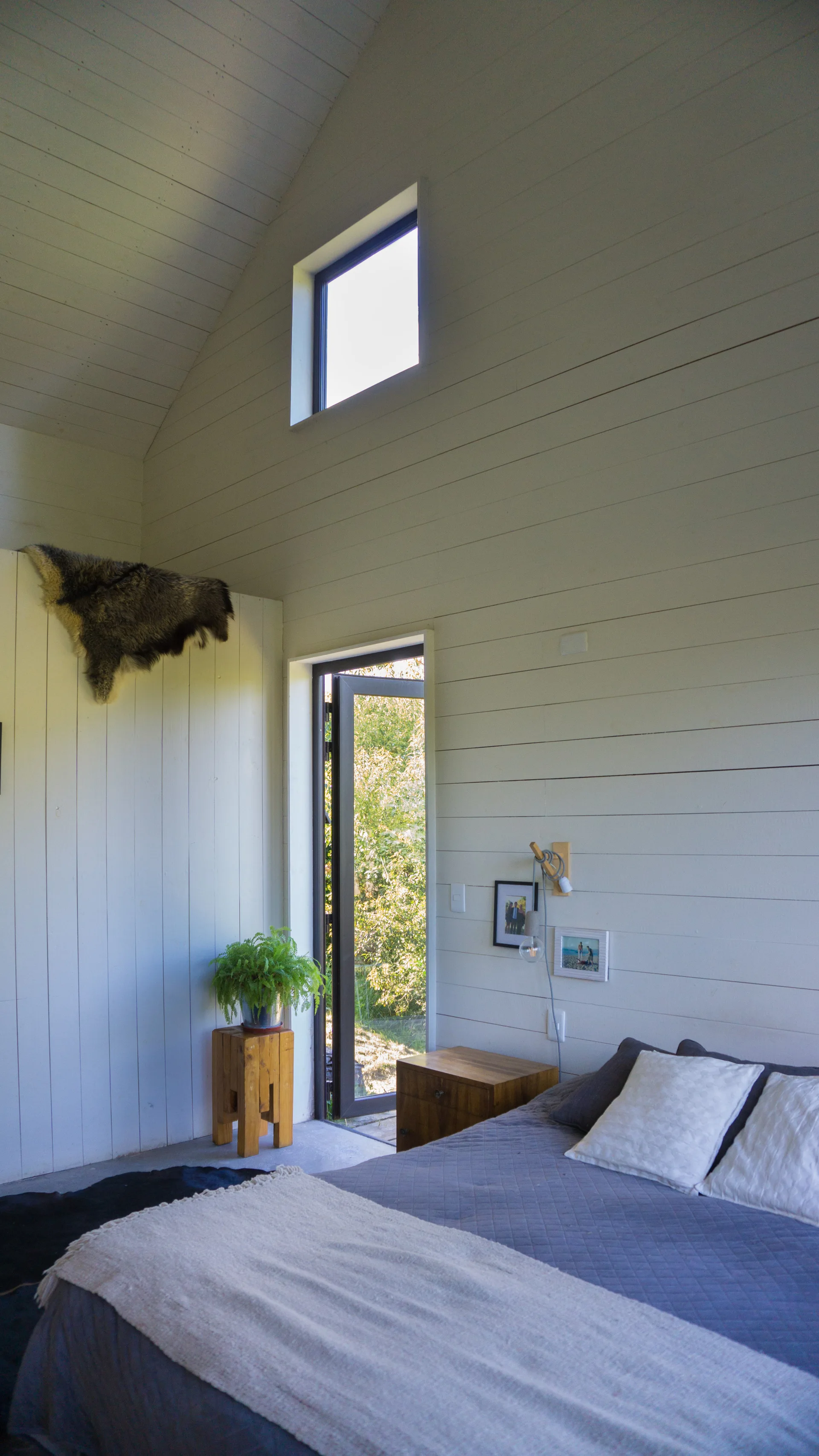 A minimalist bedroom featuring white horizontal wood paneled walls, a vaulted ceiling, a grey quilted bed, and a glass door opening to the outdoors.