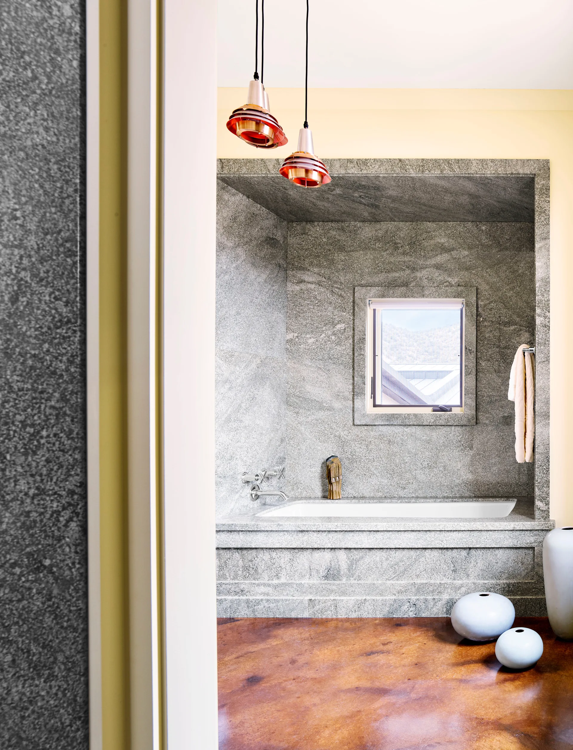 A built-in white bathtub is nestled inside a grey granite alcove with a small square window and copper pendant lights hanging above.