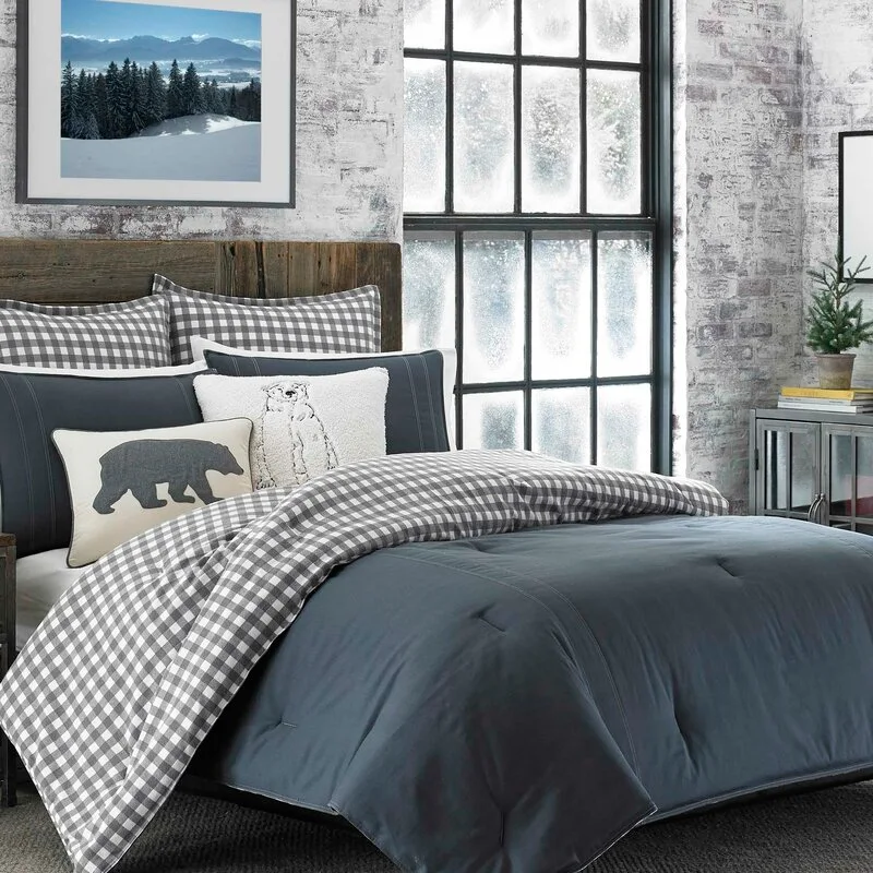 A bed dressed in navy blue and grey gingham bedding stands against a white painted exposed brick wall next to a large black grid window.
