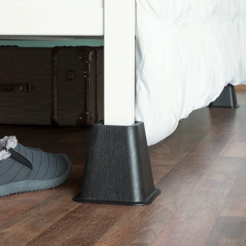 A black tapered bed riser sits under a white wooden bed leg on dark wood plank flooring next to a grey slipper.