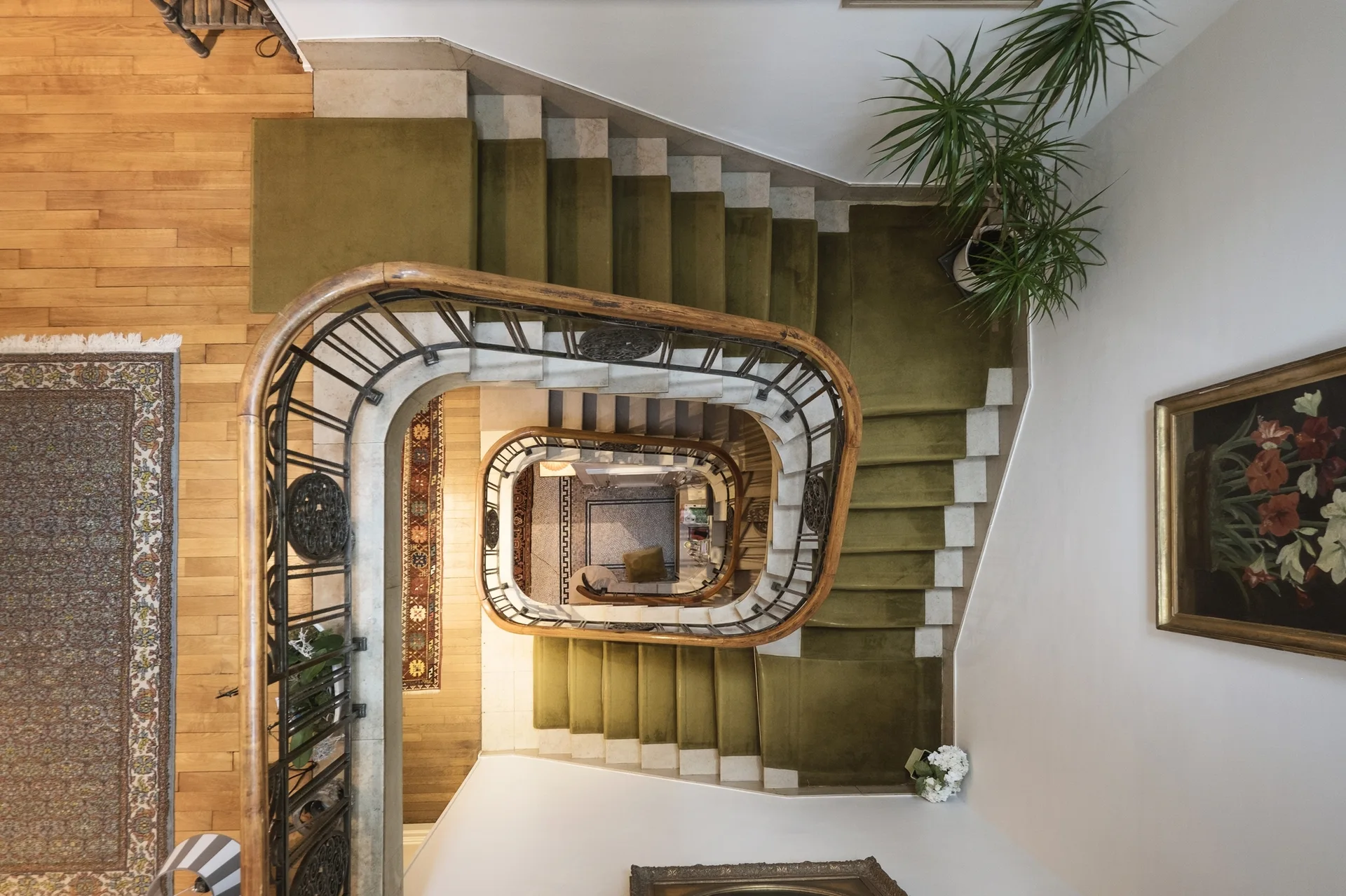 A top-down view of a curved staircase featuring an olive green carpet runner, an ornate metal railing, and a polished wood banister.
