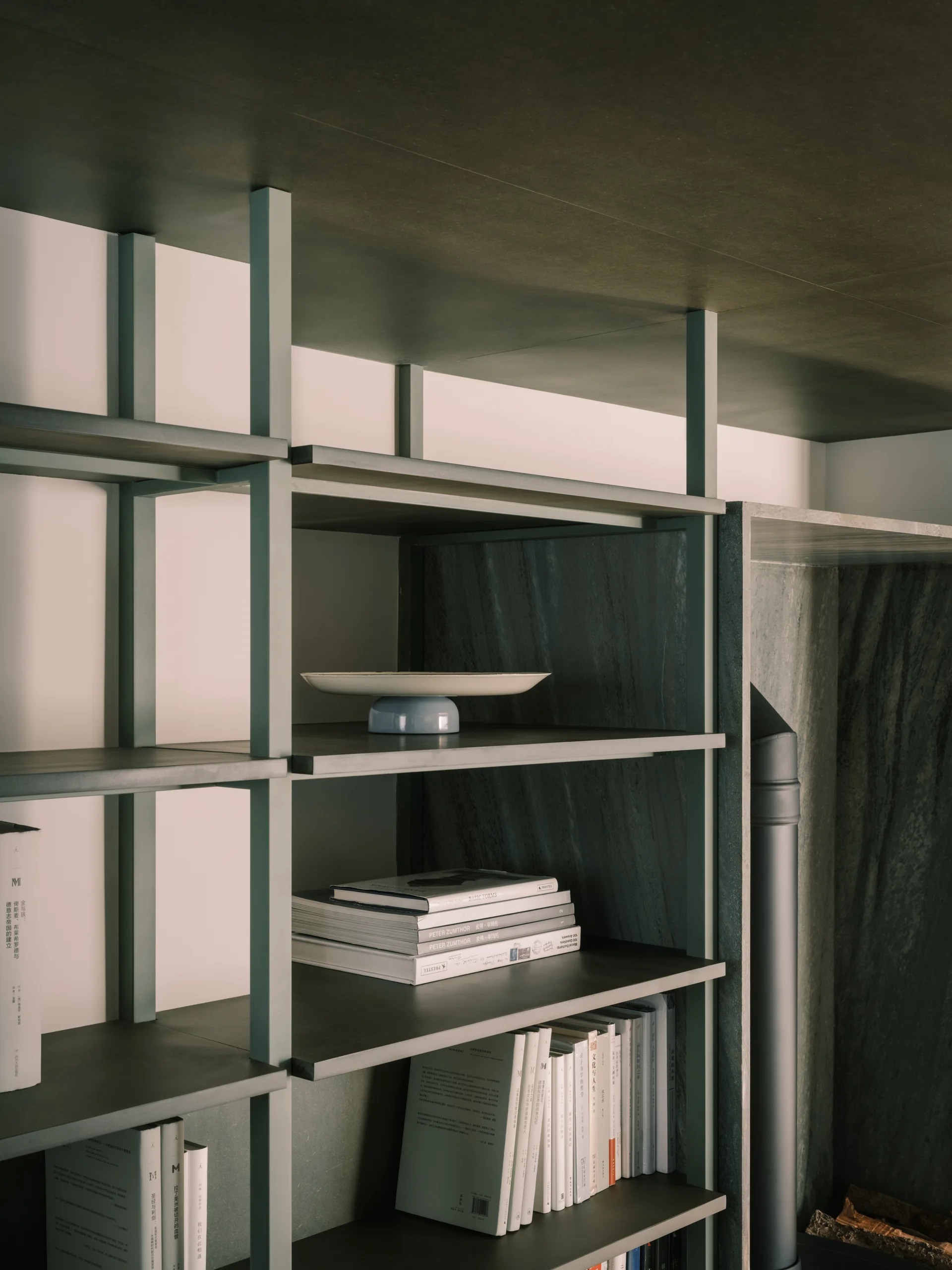 A dark grey metal open shelving unit displaying books and a ceramic dish against a dark, textured stone wall panel.
