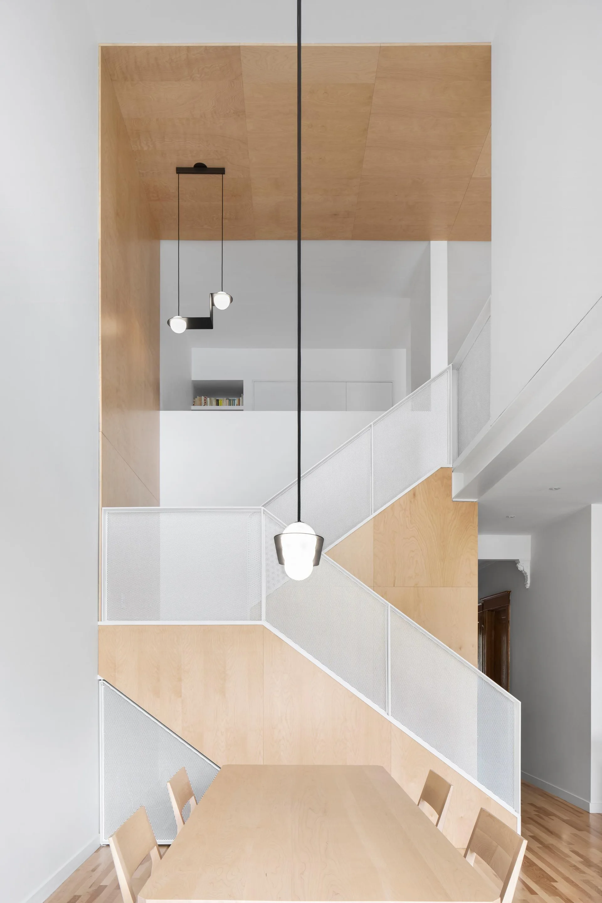 A modern staircase with light plywood wall panels, a white perforated metal mesh railing, and black pendant lights hanging over a wood dining table.
