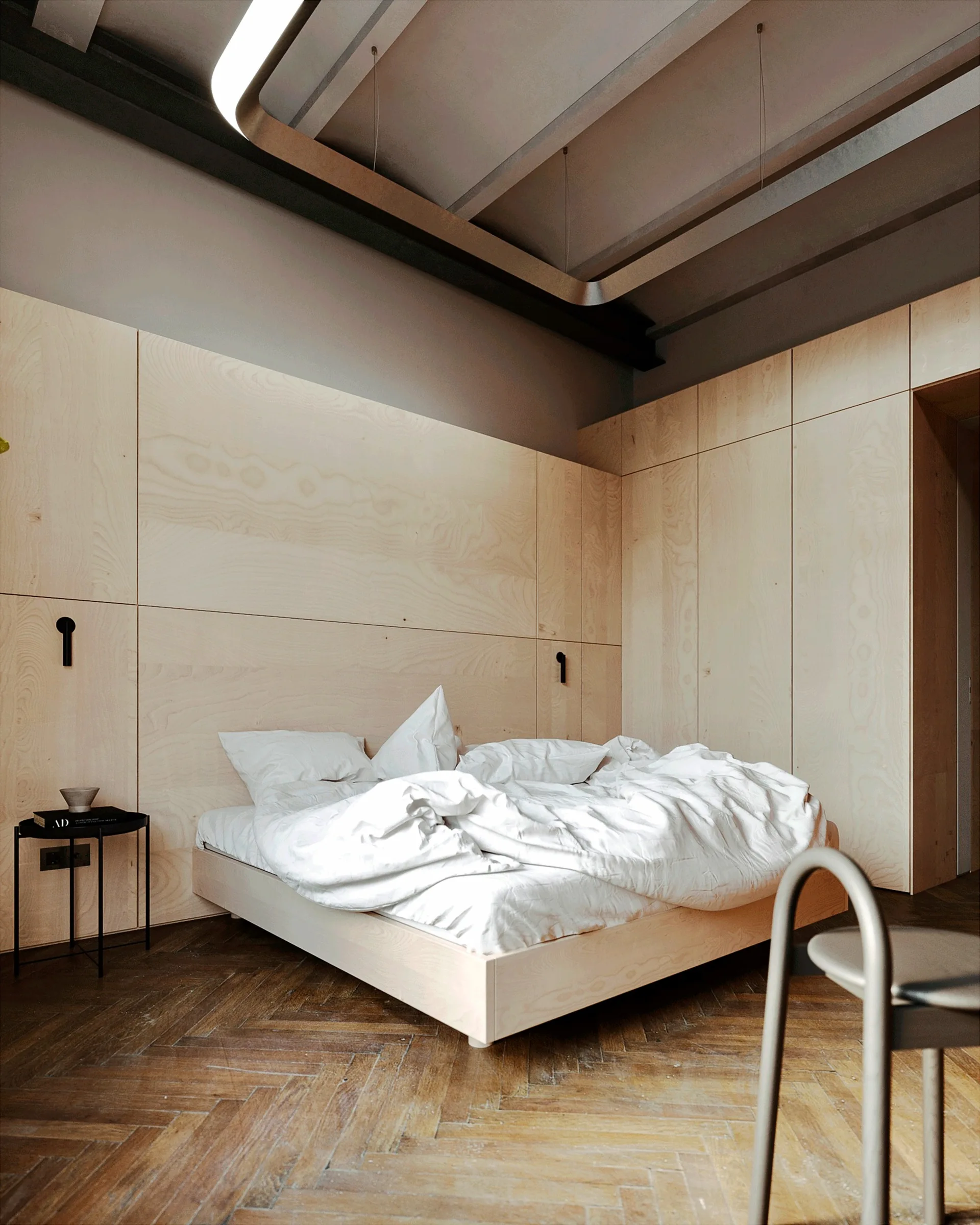 A minimalist bedroom featuring a low wooden platform bed set against light plywood wall paneling and dark herringbone parquet flooring.