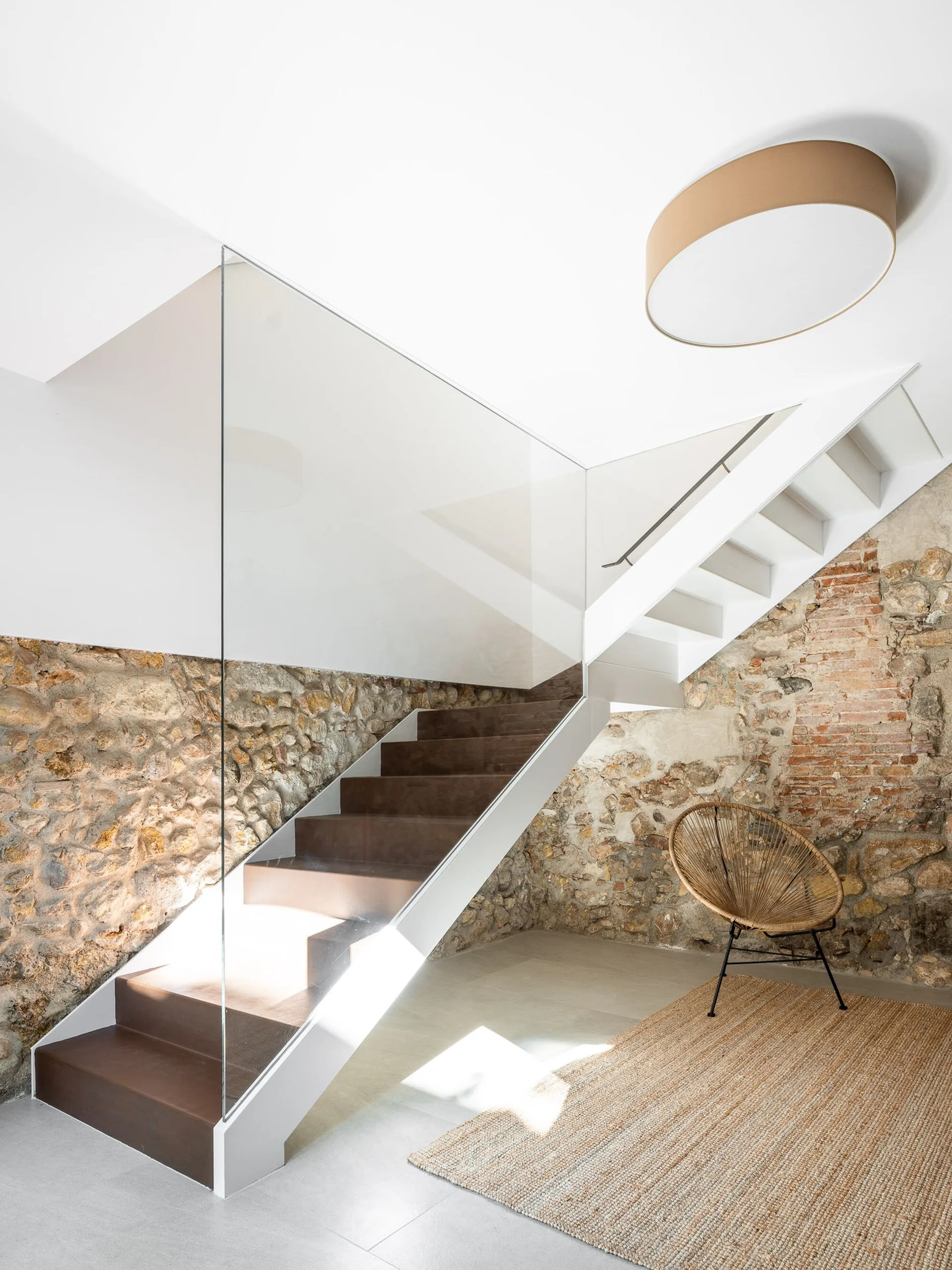 A modern white staircase with a frameless glass railing rises against an old exposed stone wall next to a wicker chair and woven rug.
