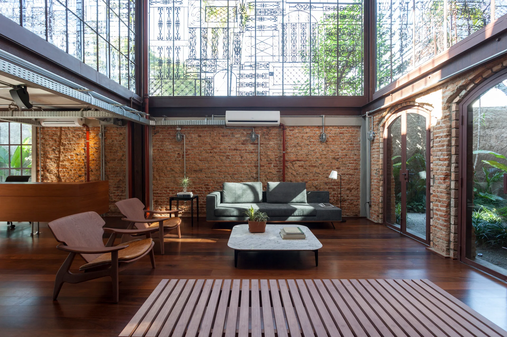 An industrial living room featuring a grey sofa, wooden armchairs, a marble coffee table, exposed brick walls, and tall ornate metal-framed windows.