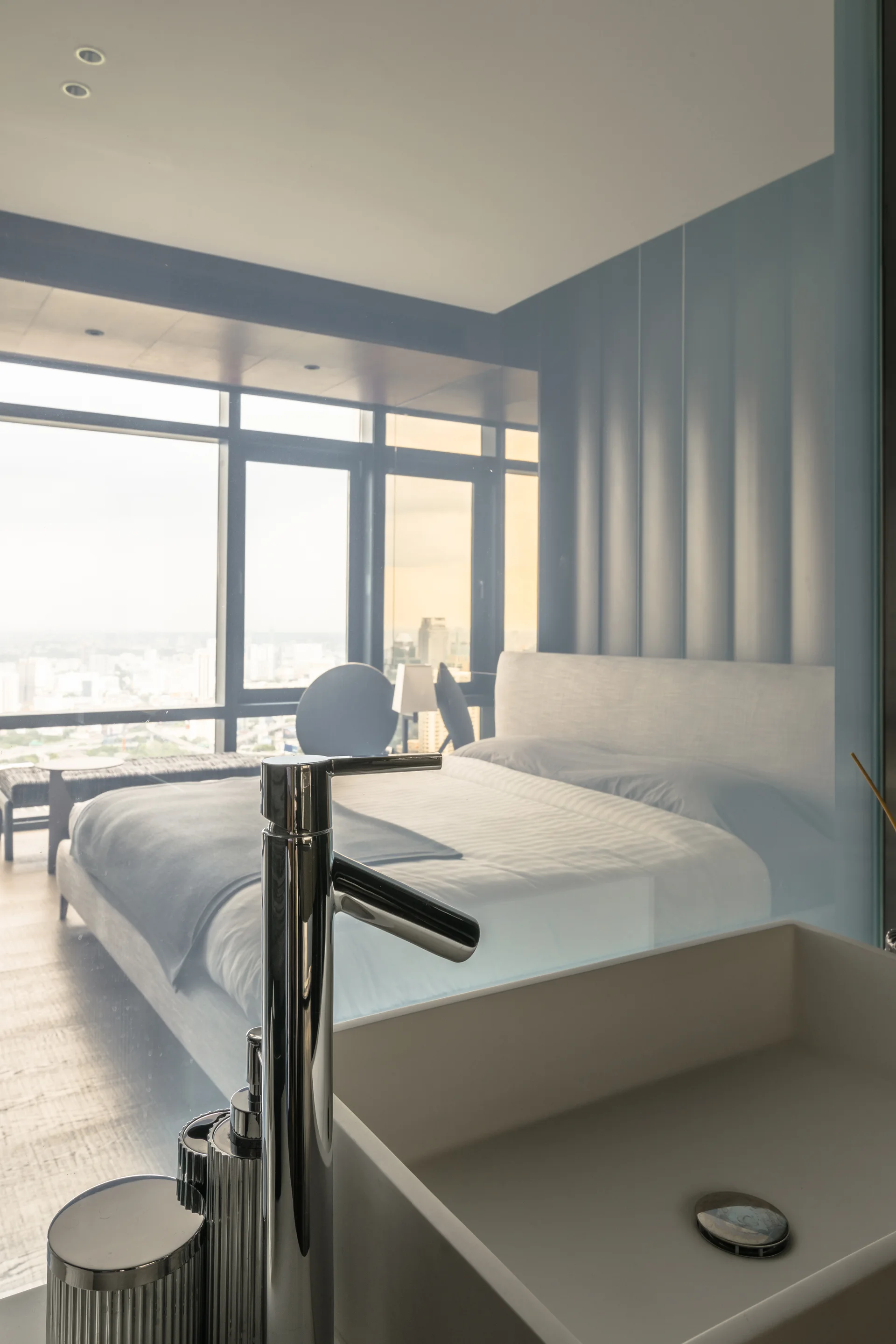 A white rectangular washbasin and chrome faucet in the foreground look through a glass partition into a modern bedroom with a large window.