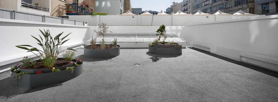 Large circular dark gray metal planters containing green plants and small trees sit on a gray paved rooftop terrace with white walls.