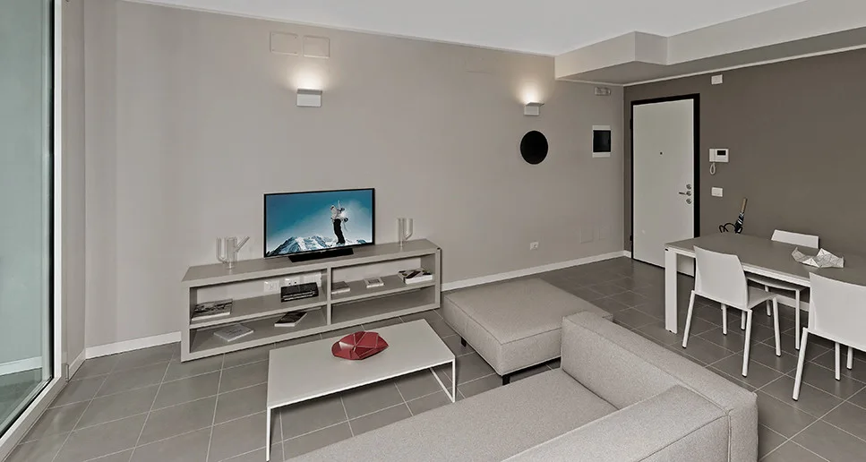 A minimalist living room features a grey tiled floor, a light grey sectional sofa, a low media console with a TV, and a dining table with white chairs.