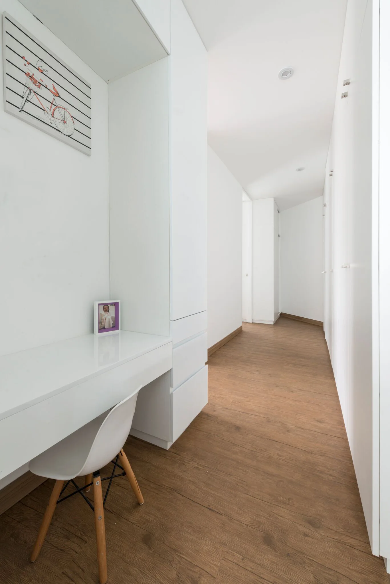 A minimalist hallway features a white built-in desk with a white chair on brown wood floors next to floor-to-ceiling white cabinets.