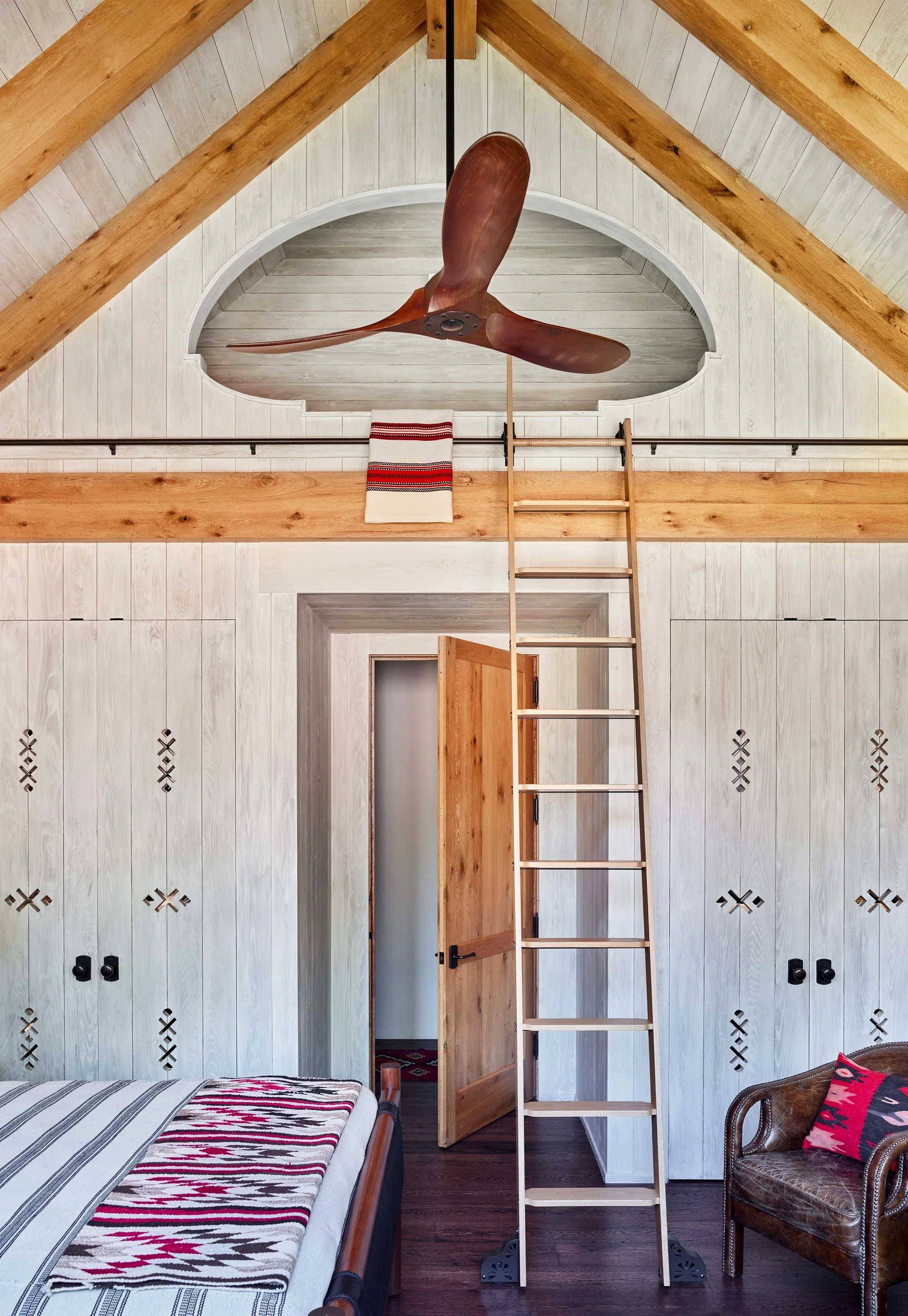 A cabin bedroom featuring whitewashed wood paneling, a vaulted ceiling with exposed timber beams, a ceiling fan, and a rolling wooden ladder.