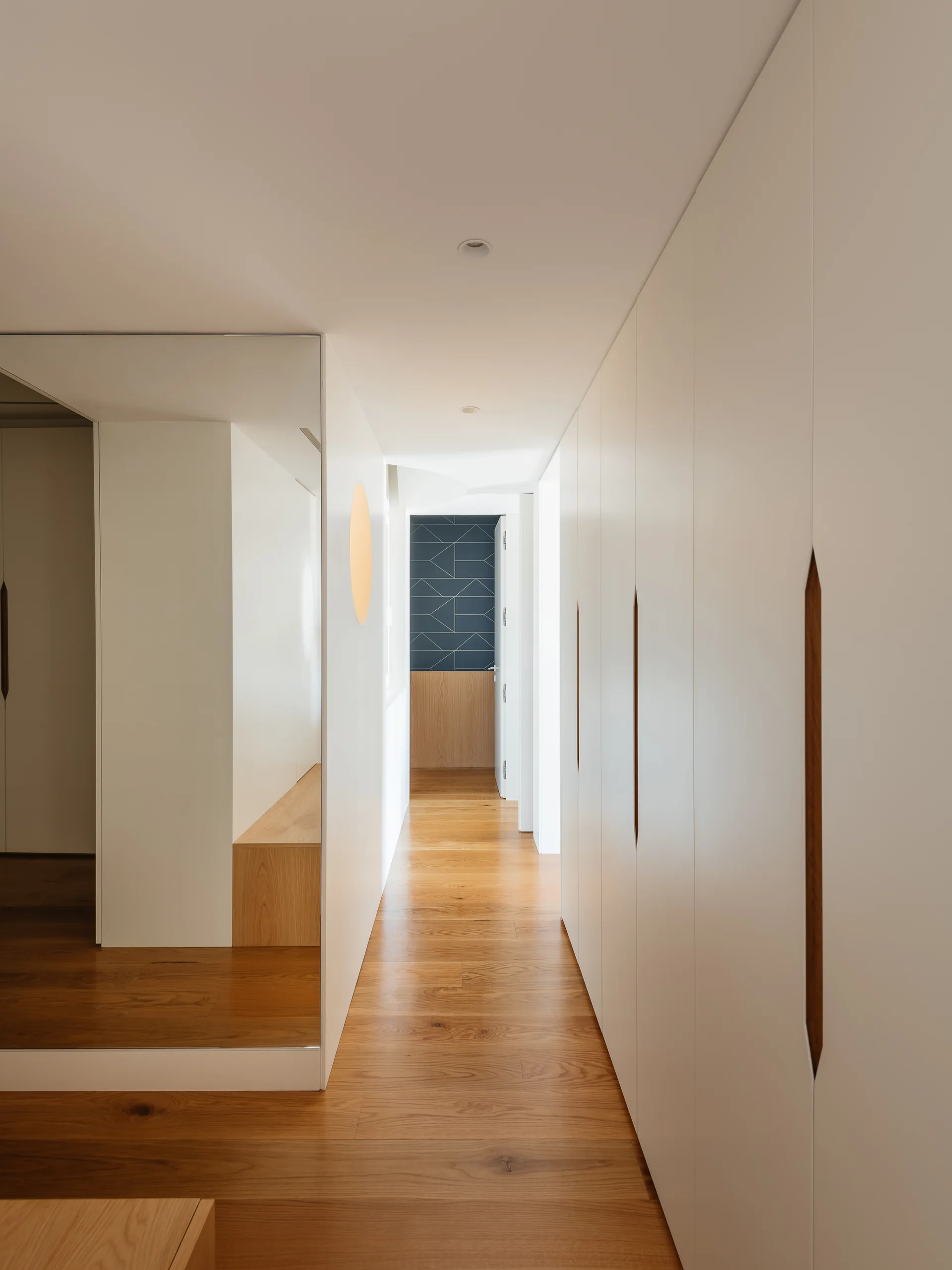 A narrow white hallway with built-in storage cabinets featuring carved wood handles, a large wall mirror, and light hardwood floors.