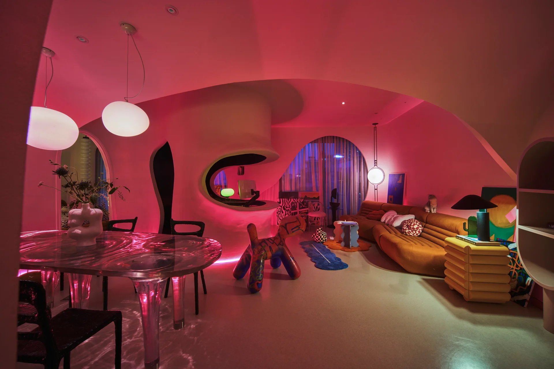 An organic living and dining area with curved pink plaster walls, a translucent ripple-textured table, and a low tan sofa.