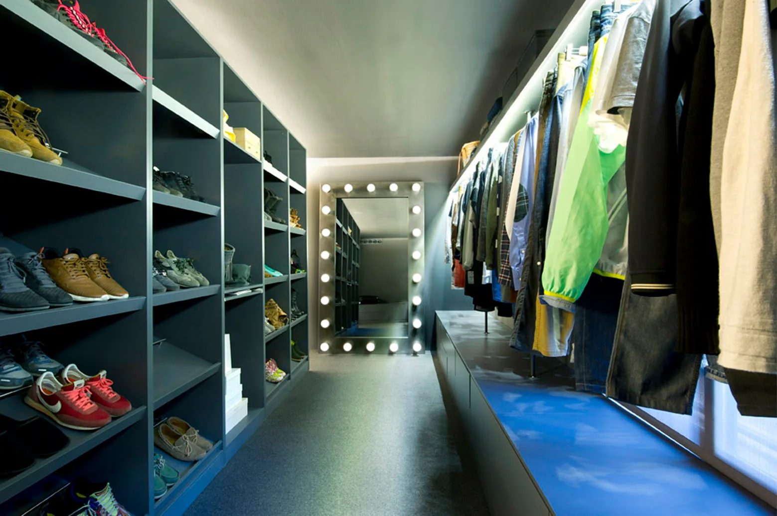 A narrow walk-in closet with dark gray shoe shelving, hanging clothing, and a large lighted vanity mirror at the end of the aisle.