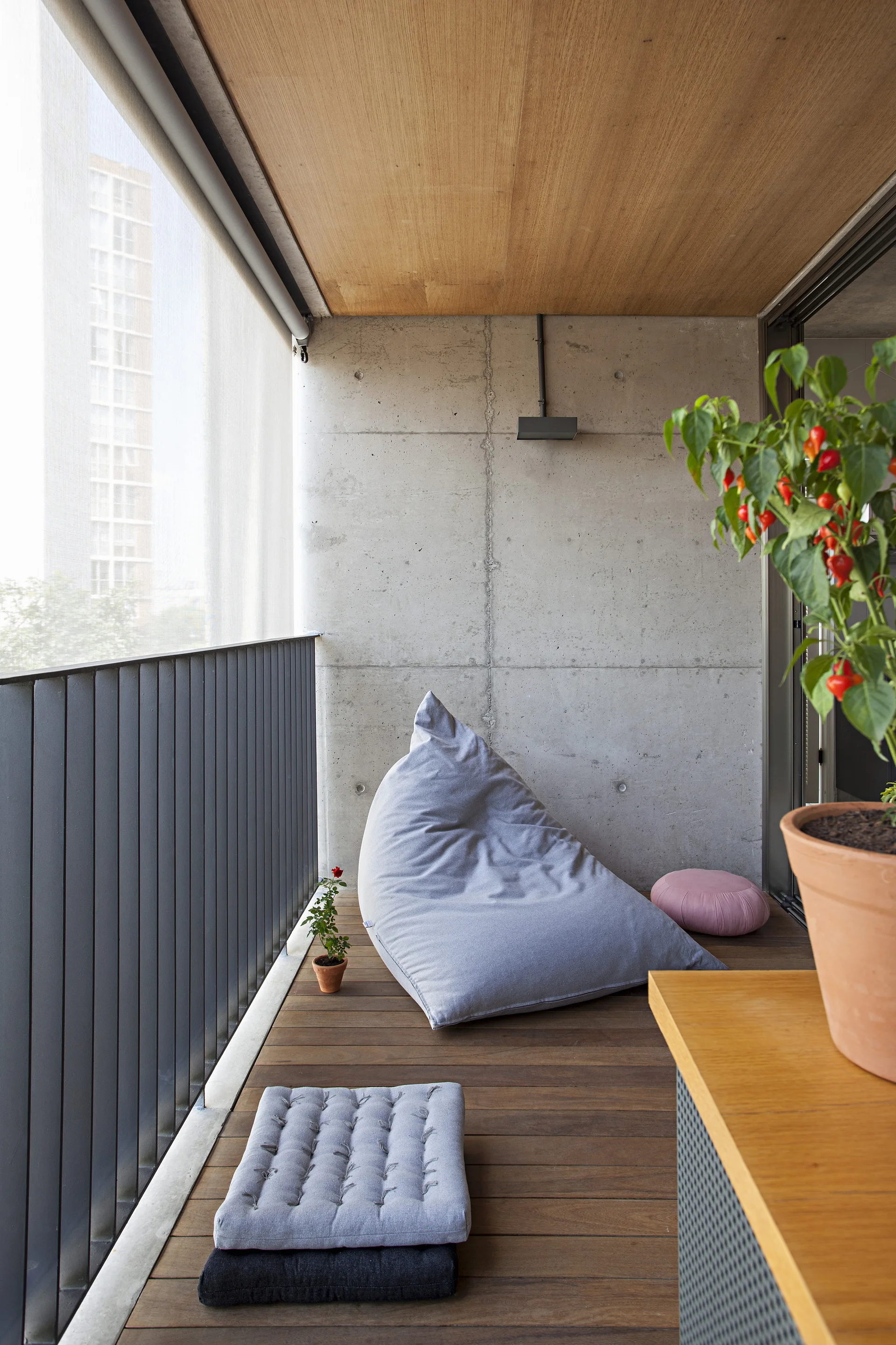 A grey bean bag chair and floor cushions sit on a wood-decked balcony with an exposed concrete wall and wood ceiling panel.