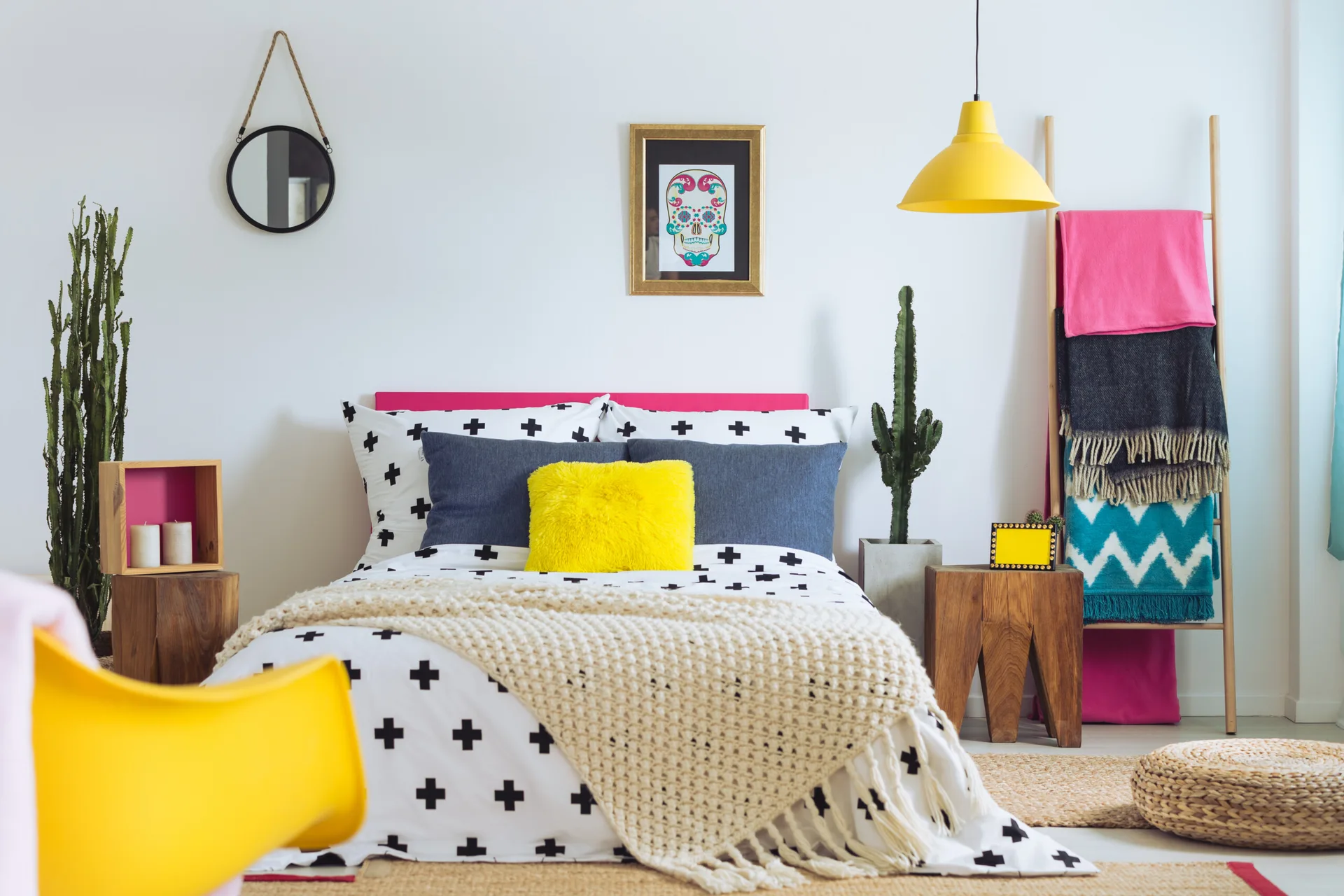 An eclectic bedroom features a bed with graphic pattern bedding, a pink headboard, blocky wooden side tables, potted cacti, and a yellow pendant light.