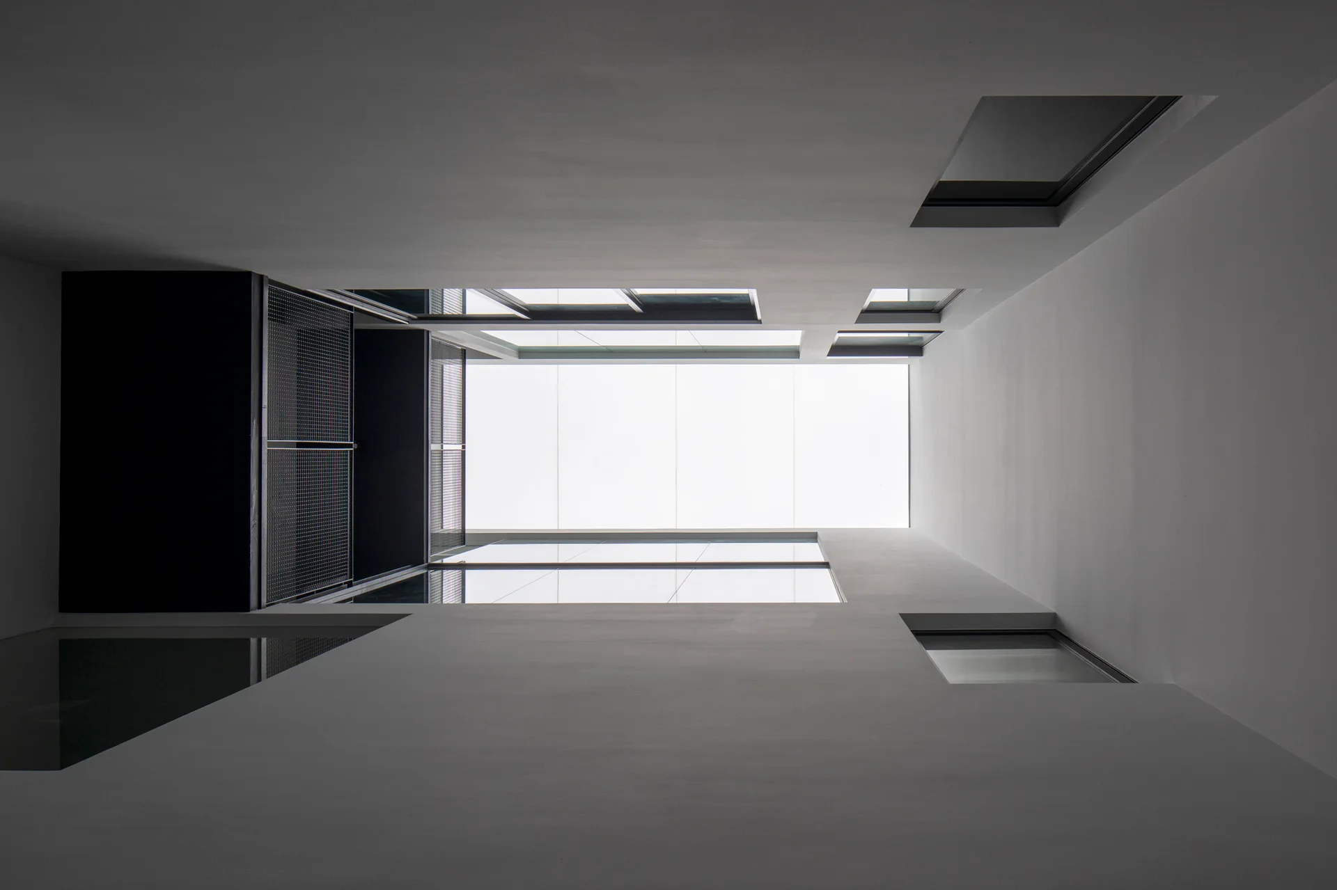 An upward look through a minimalist white interior light well featuring a bright overhead skylight and dark geometric window frames.