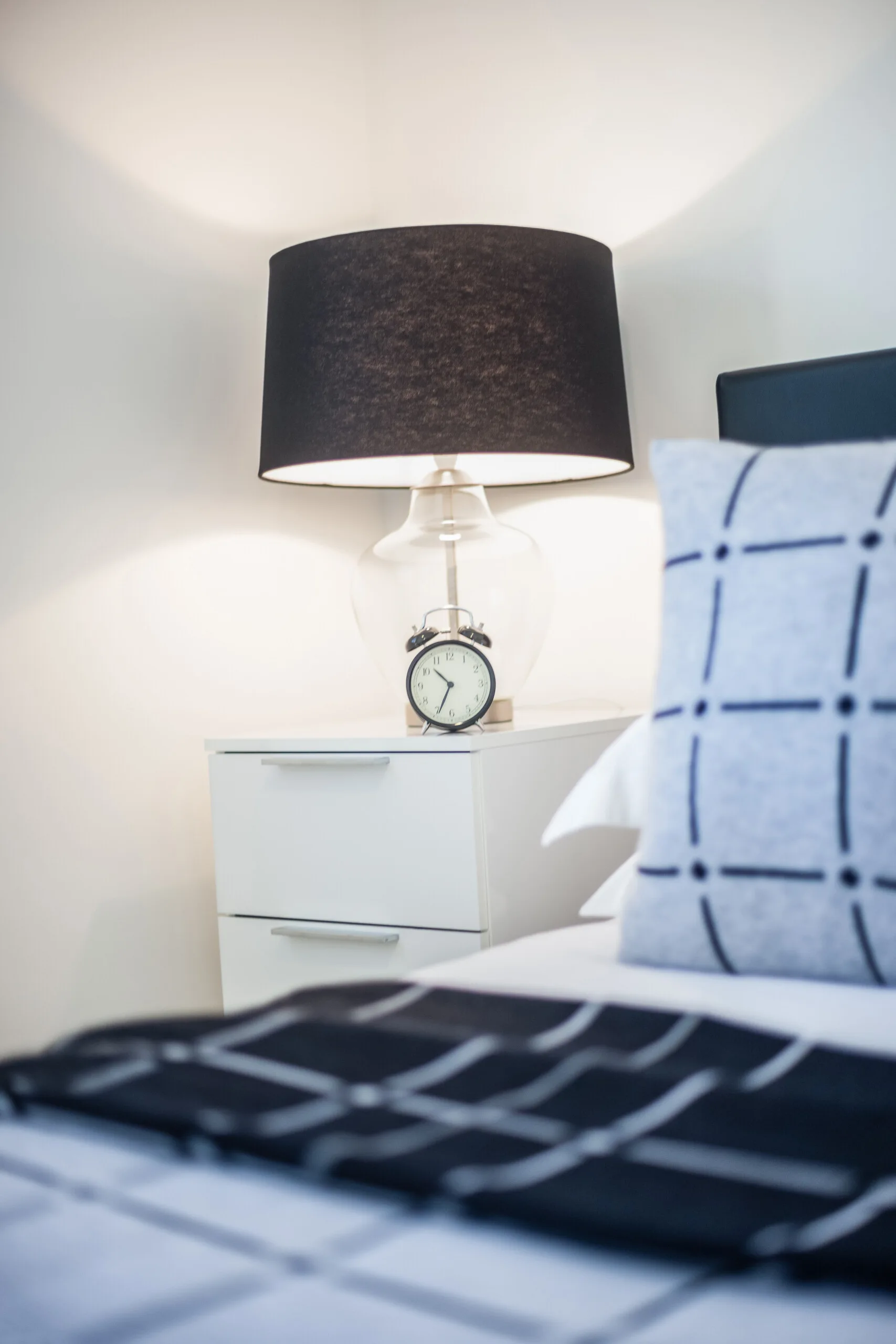 A minimalist white nightstand holds a clear glass lamp with a black shade beside a bed with grid patterned linens.