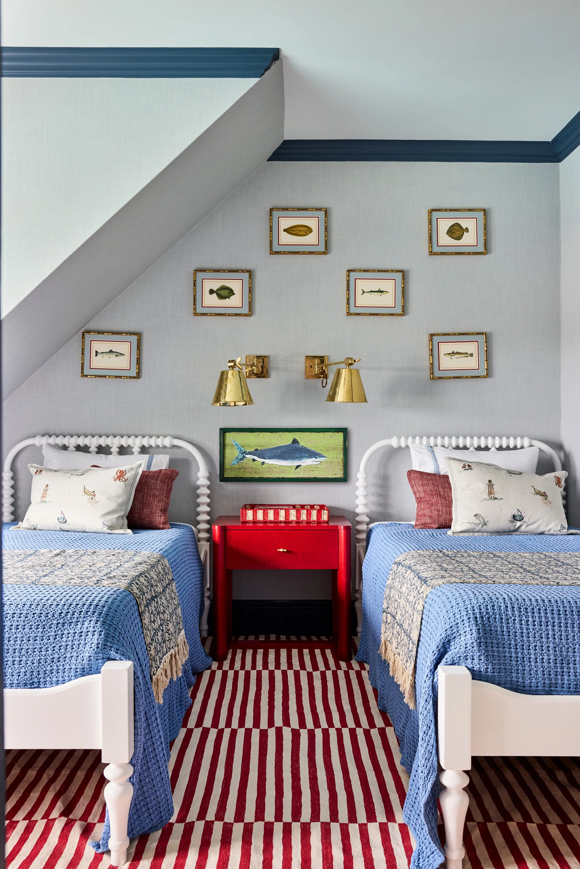 Two white spool twin beds flank a bright red nightstand beneath framed fish prints in a bedroom with a sloped ceiling and blue trim.