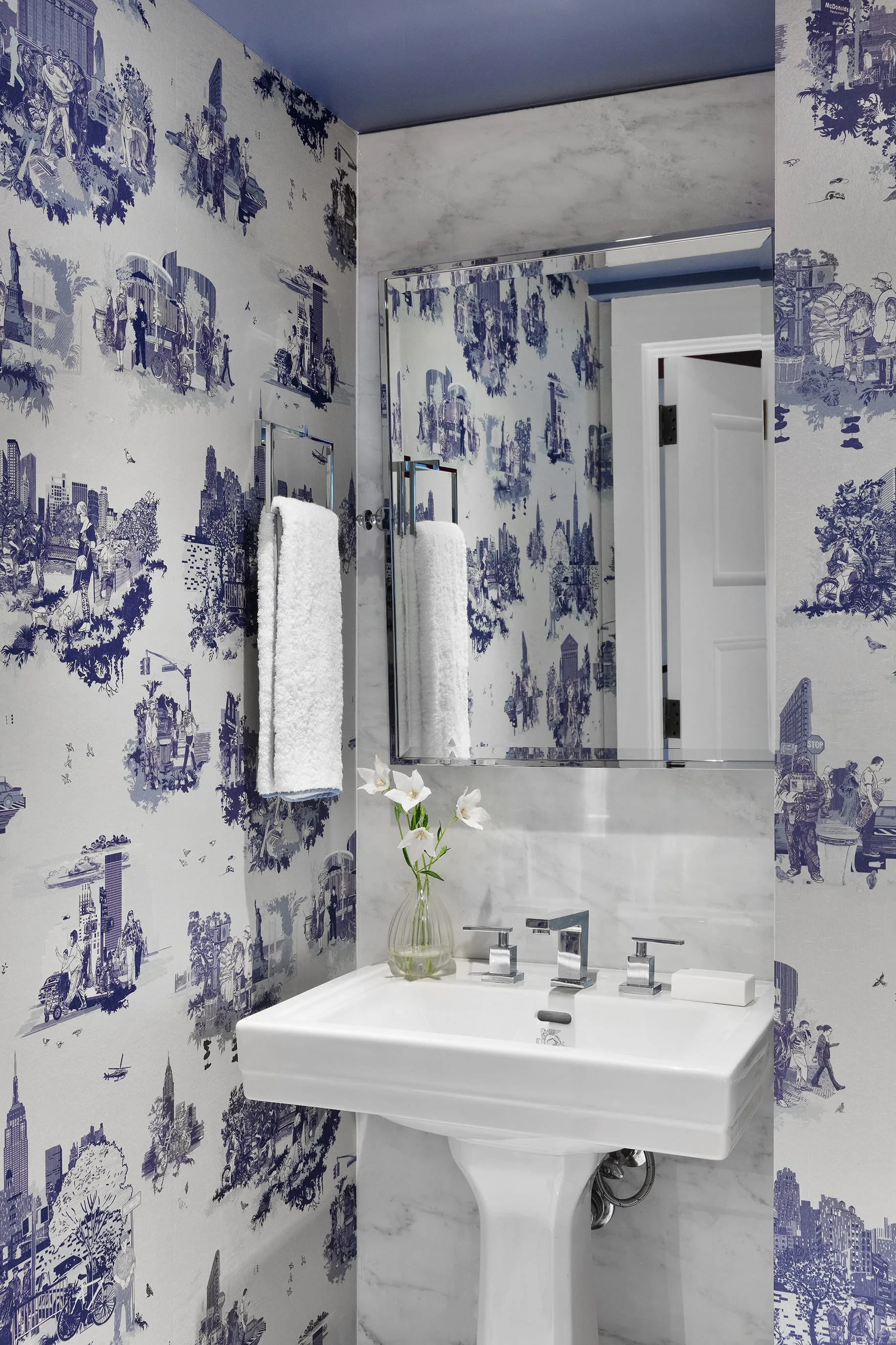 A compact powder room featuring blue and white city toile wallpaper, a white pedestal sink, a marble tiled backsplash, and a chrome-framed mirror.