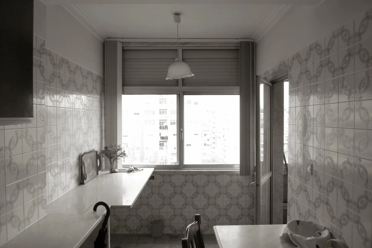 A narrow kitchen with white patterned wall tiles, a long countertop, a central window, and a hanging pendant light.