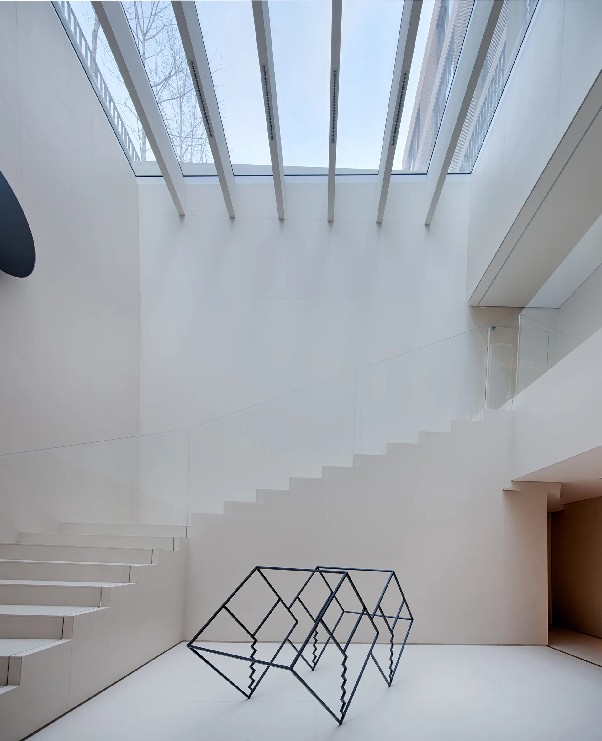 A modern white atrium features a minimalist staircase with a glass railing, a black geometric sculpture on the floor, and an overhead glass skylight.