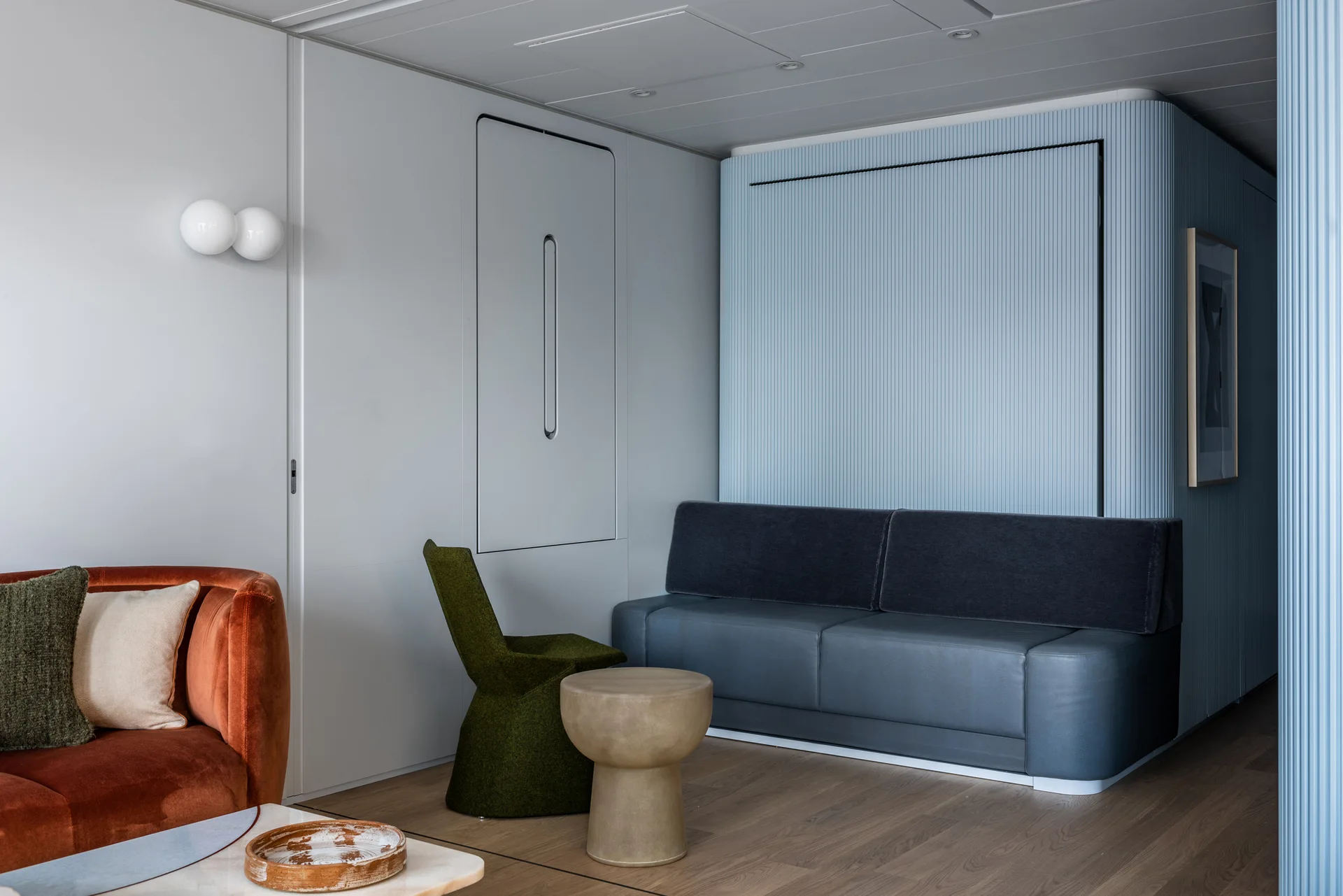 A contemporary living room featuring a pale blue fluted wall behind a grey sofa, an orange velvet sofa, and a sculptural green chair on hardwood floors.
