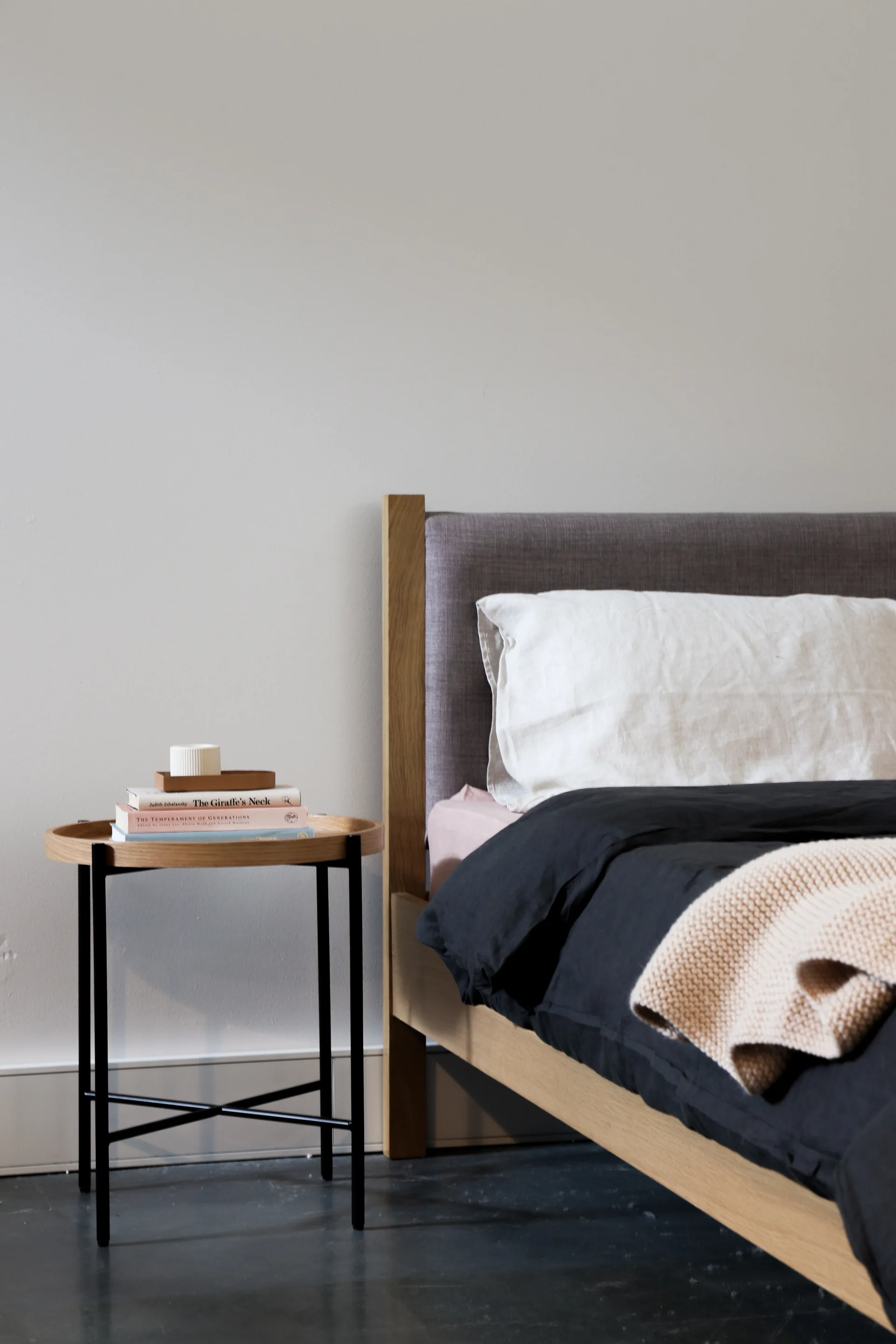 A modern wood bed frame with a grey headboard sits beside a round tray-top nightstand holding books against an off-white wall.