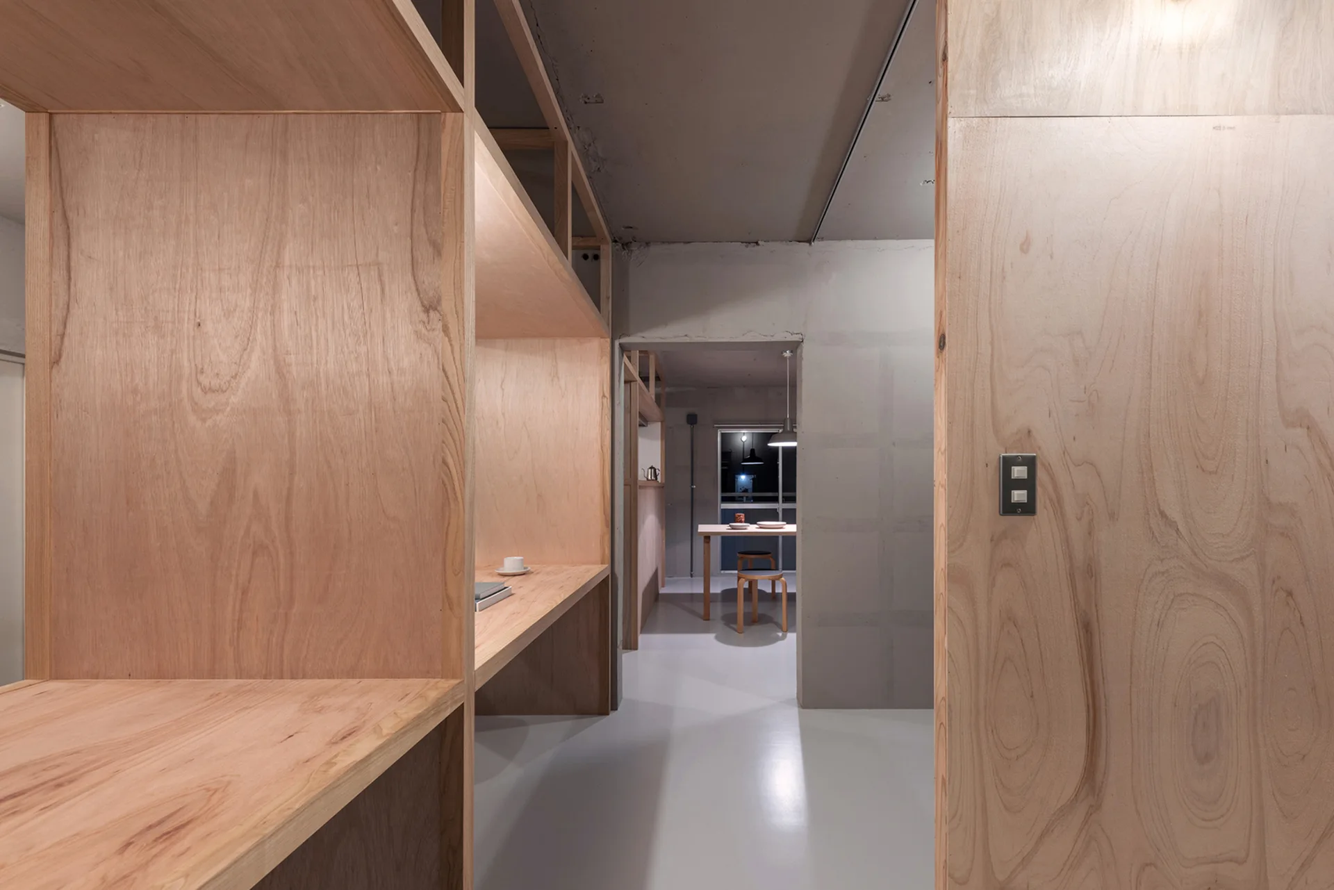 Minimalist room featuring light-toned plywood built-in shelving, raw concrete walls, and a view into a dining area with a wooden table.