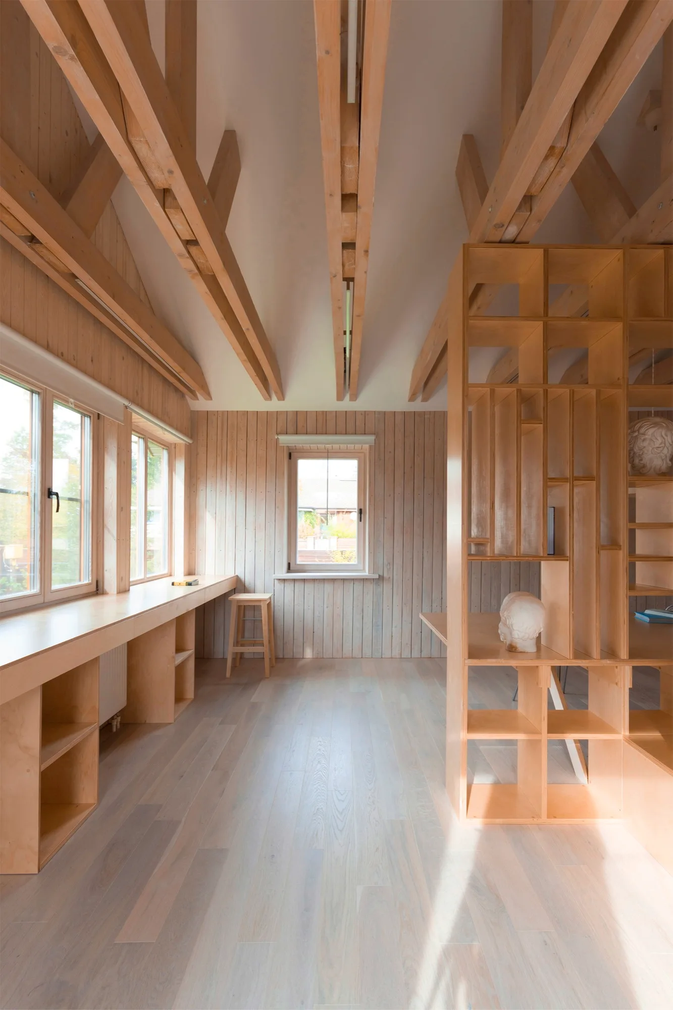 A minimalist home office featuring light wood wall paneling, a long built-in plywood desk below multiple windows, and exposed timber ceiling beams.