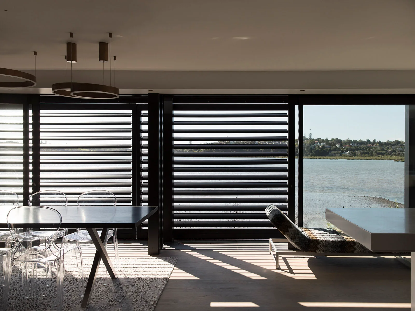 A modern dining room features a dark table with translucent plastic chairs set against large windows with black horizontal shutters overlooking the water.