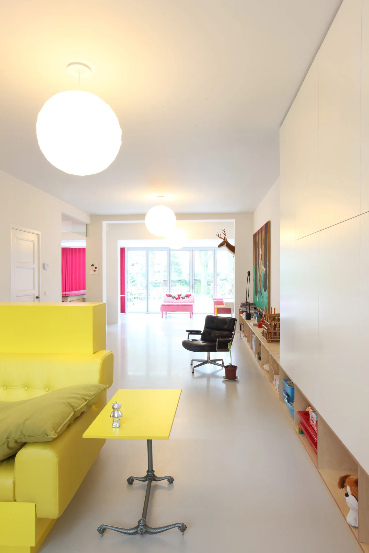 A minimalist living room with a bright yellow sofa, spherical ceiling lights, and integrated white wall cabinets with open base shelves.