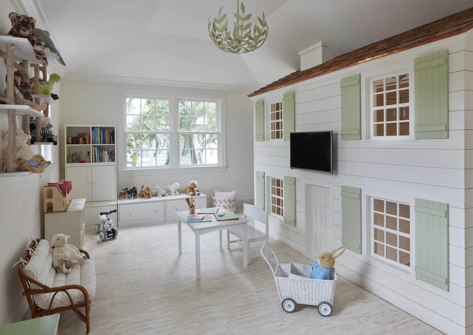 A children's playroom features a large white shiplap playhouse with sage green shutters, a small activity table, and low storage benches beneath a window.