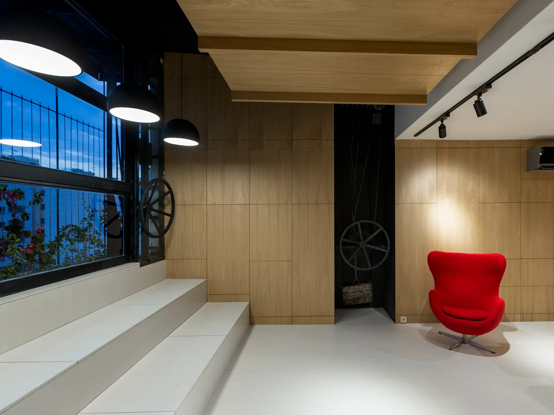 A modern lounge room featuring light wood paneled walls, a white tiered seating platform, and a bright red armchair.