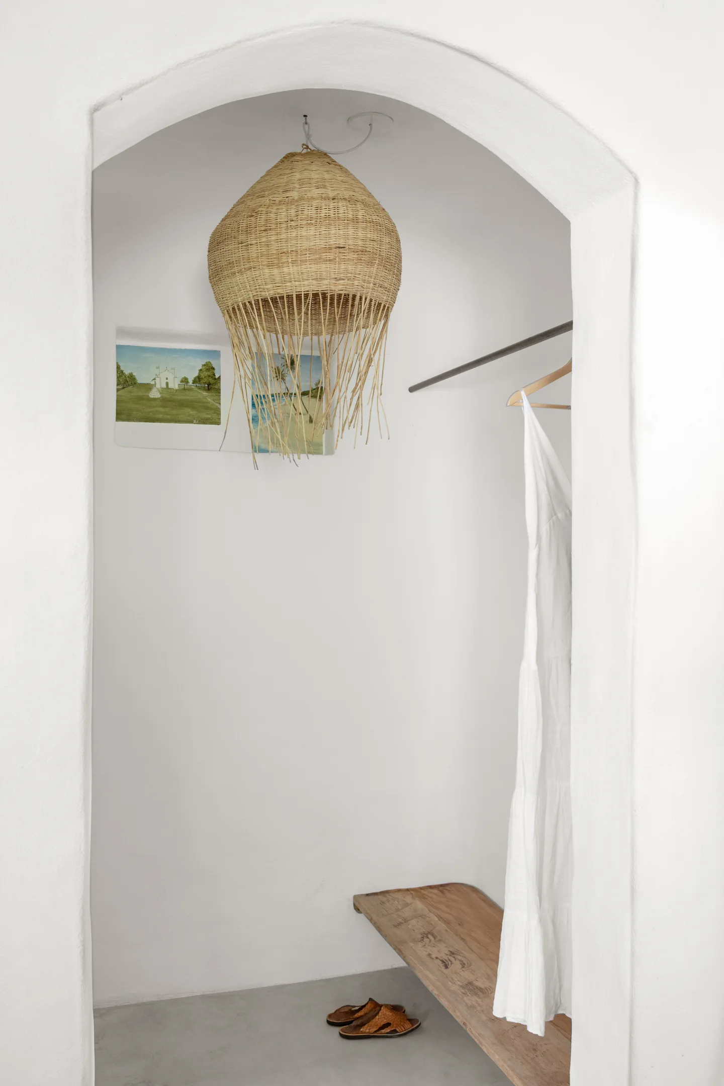 A minimalist arched closet alcove featuring white plaster walls, a wooden bench, a metal hanging rail, and a fringed woven pendant light.