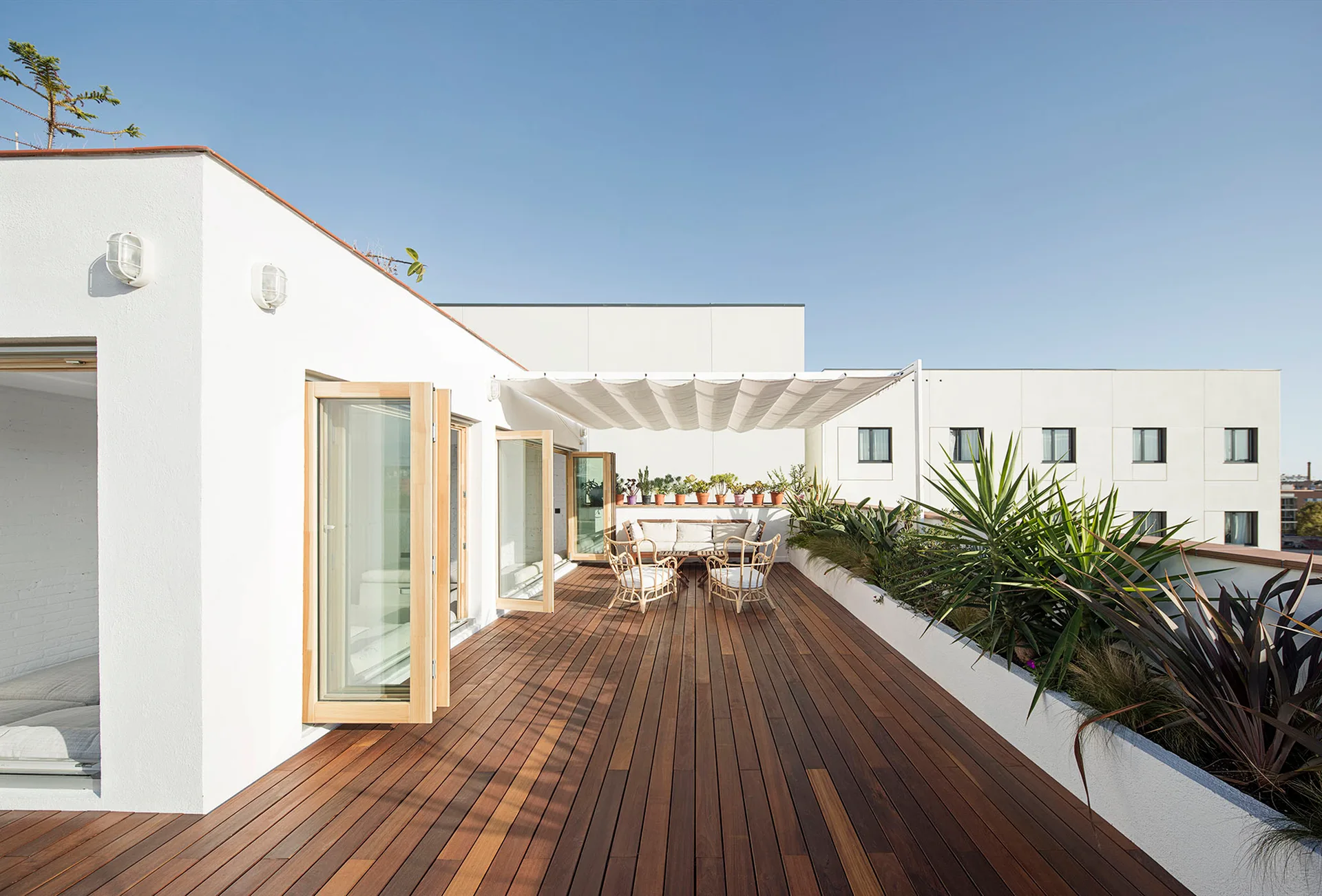 A sunlit rooftop terrace features natural wood plank decking, bi-fold glass doors, rattan outdoor furniture, and a built-in planter.