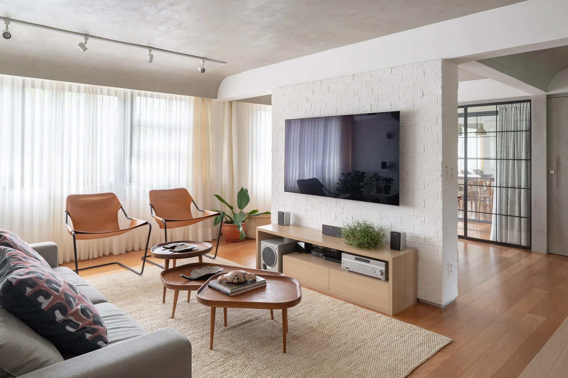 A modern living room features a grey sofa, tan leather chairs, three nested wooden coffee tables, and a white brick partition wall with a mounted TV.