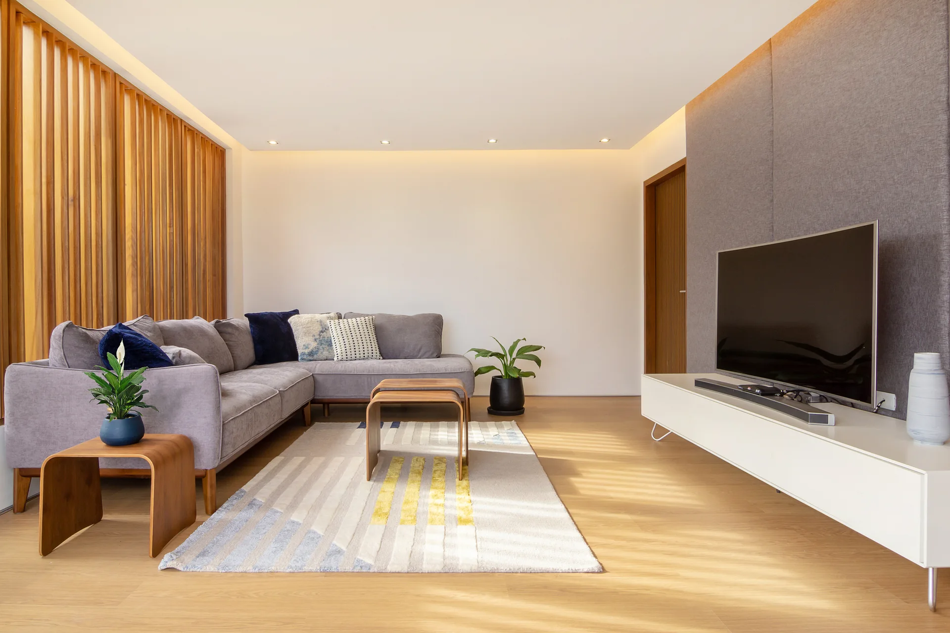A modern living room features a grey sectional sofa, curved wooden nesting tables, a white media console, and a vertical wood slat wall.