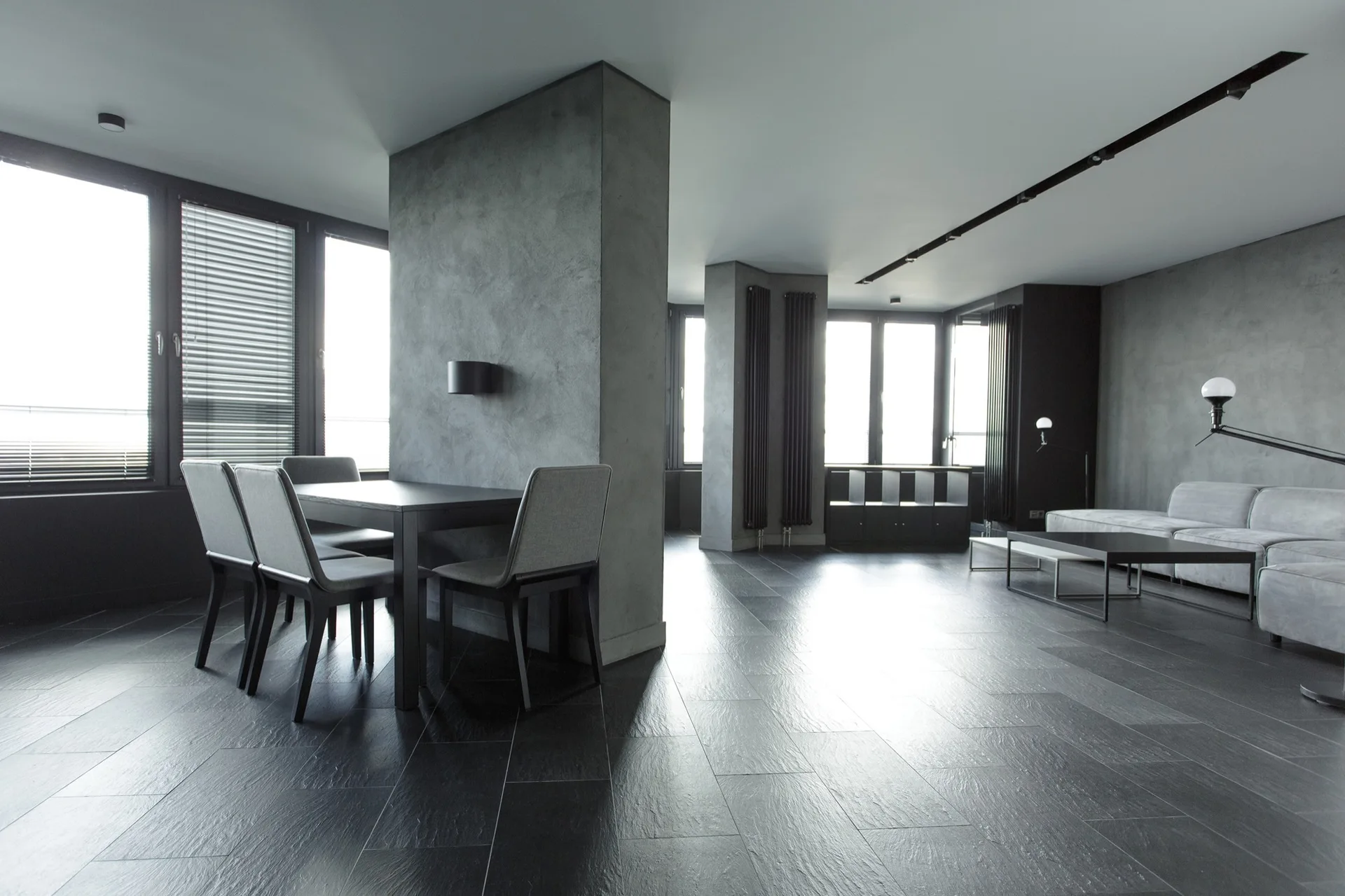 A minimalist open-plan grey apartment features a dining table next to a textured concrete column and a living area with a grey sofa.