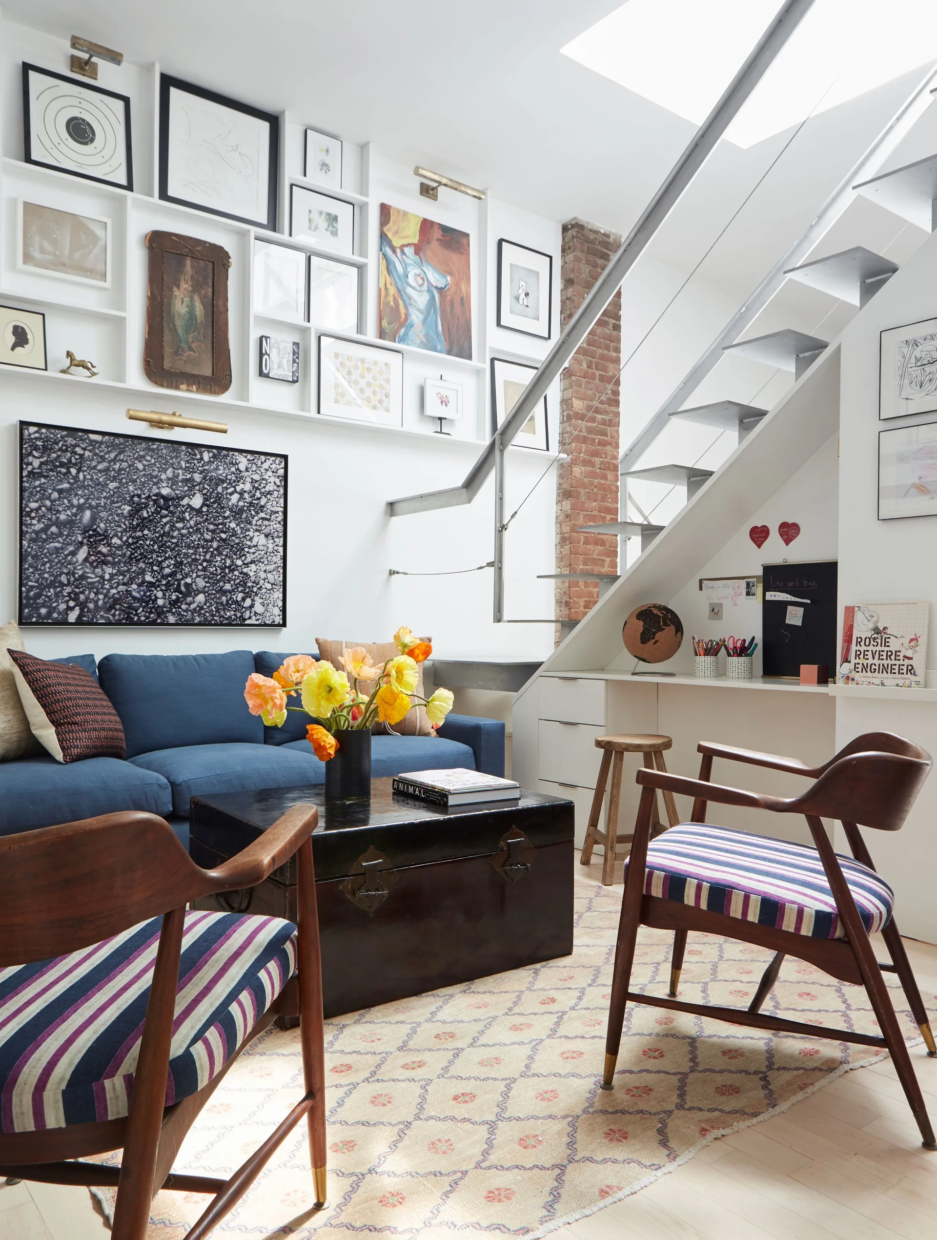 An eclectic living room features a blue sofa, striped armchairs, a trunk coffee table, and an open metal staircase alongside a massive gallery wall.