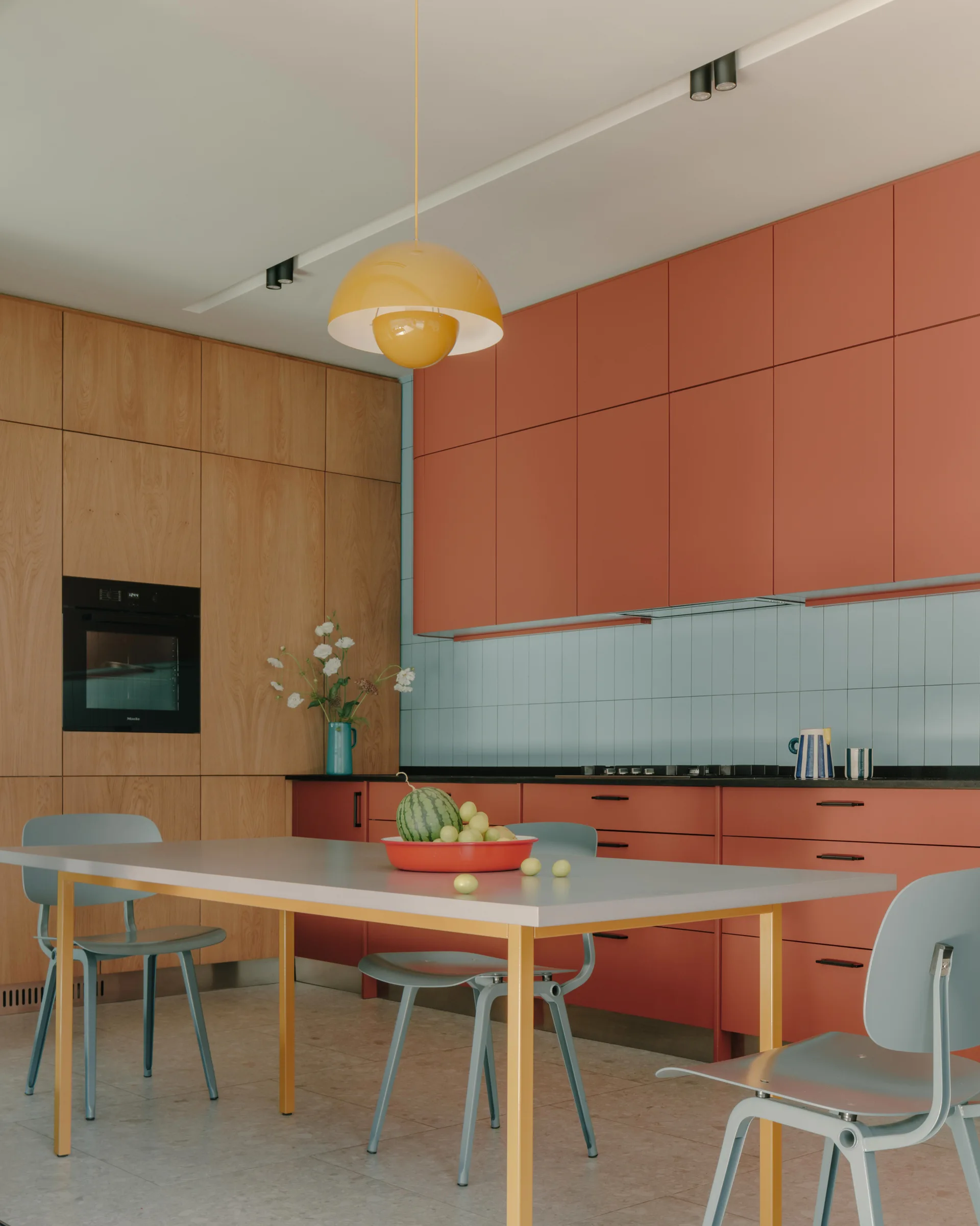 A modern kitchen featuring terra cotta cabinets, a light blue tiled backsplash, a light wood storage wall, and a gray dining table with blue chairs.