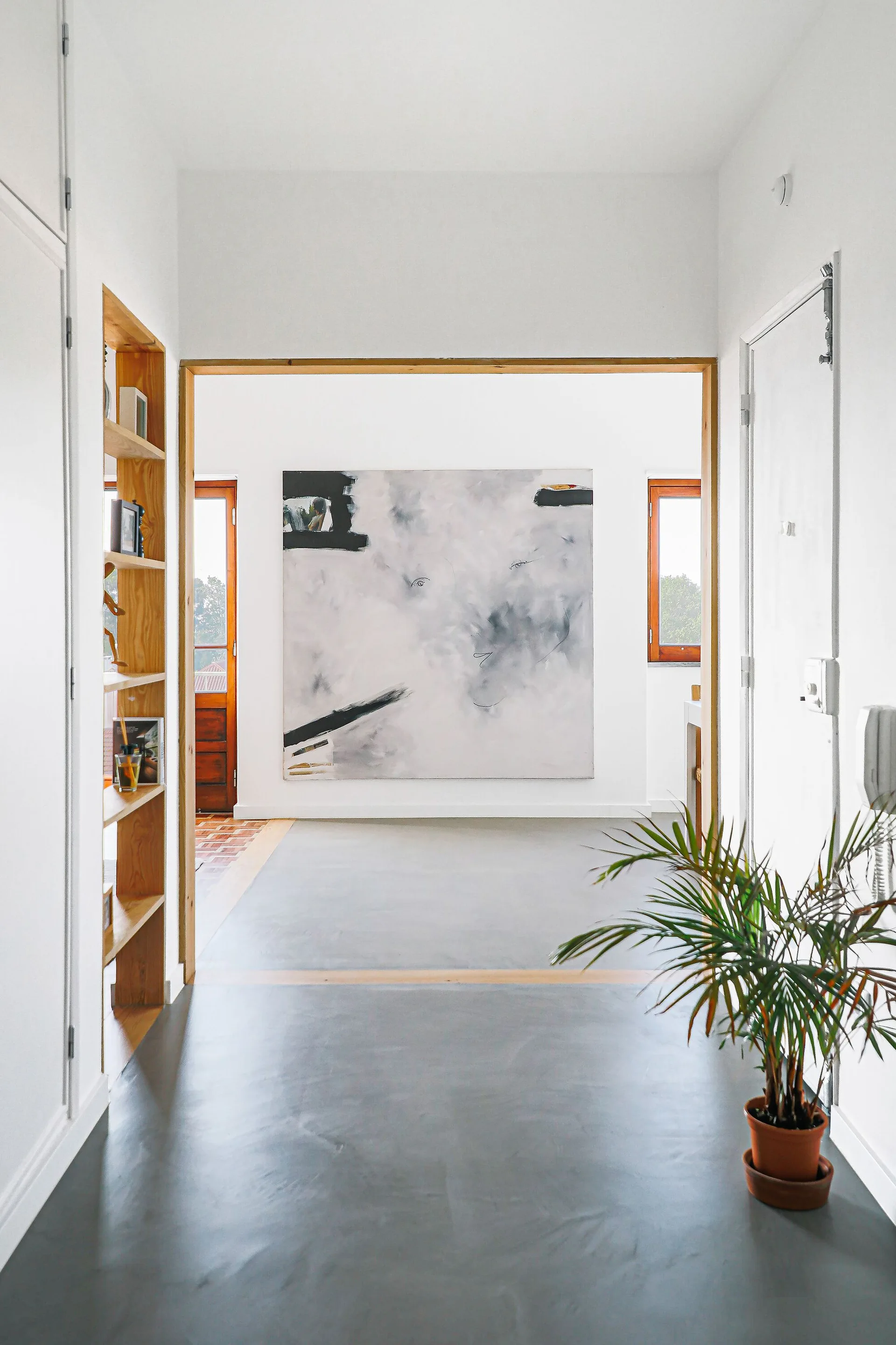 A minimalist white hallway with a grey floor features a built-in wooden bookcase, a potted plant, and a large abstract painting.