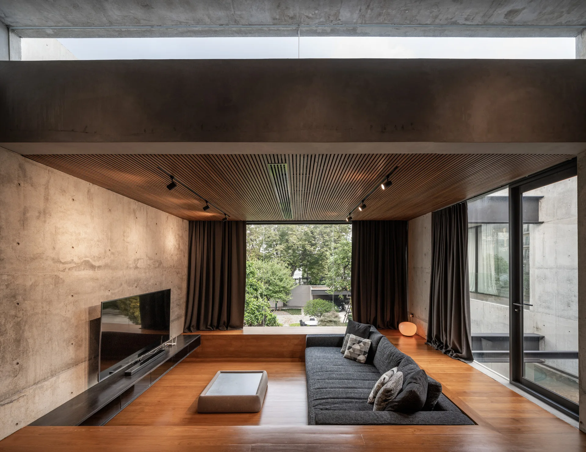 A sunken living room featuring a charcoal grey sofa recessed into wood floors, set against an exposed concrete wall and wood slat ceiling.